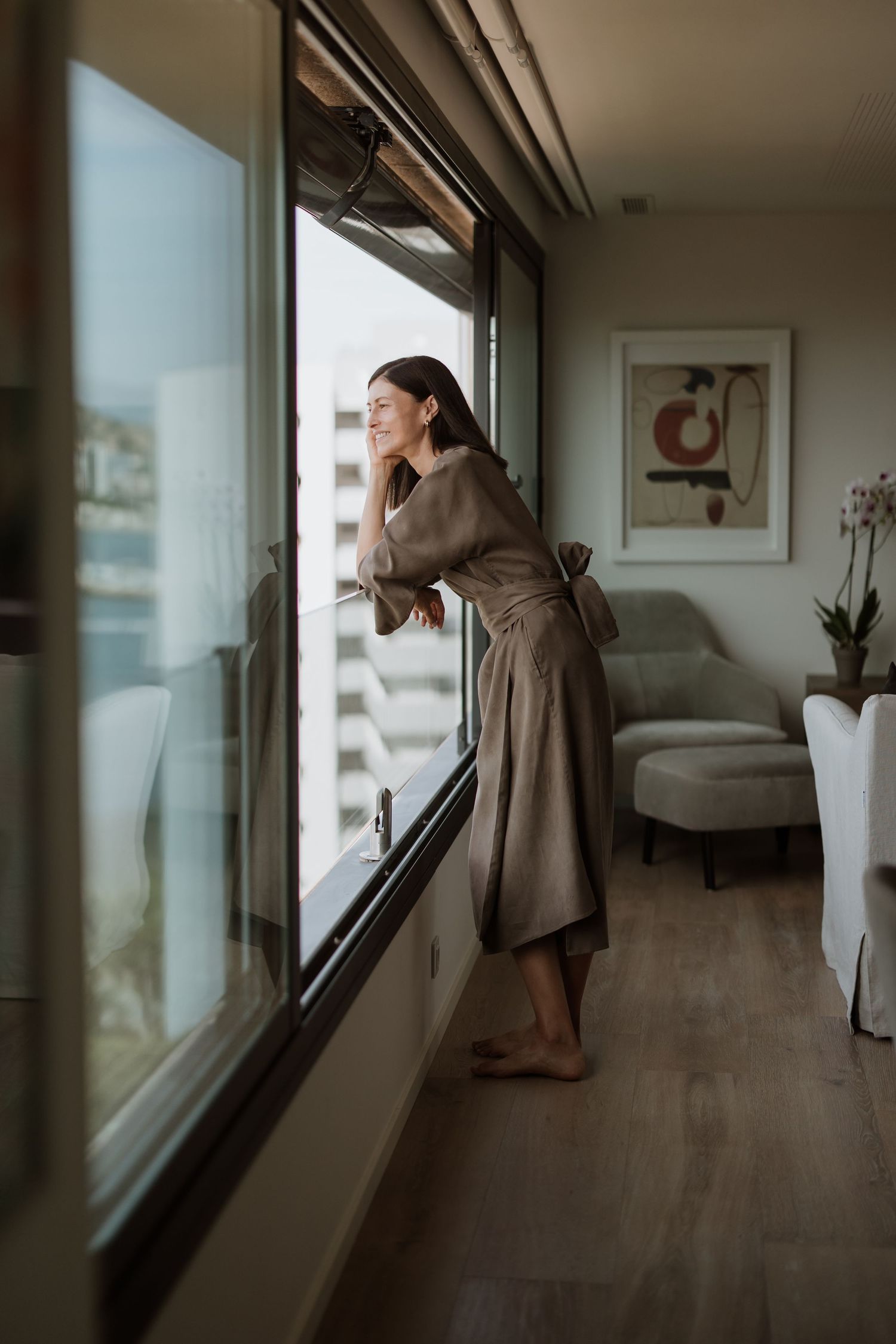 Person in brown dress looking through floor-to-ceiling windows in modern apartment.