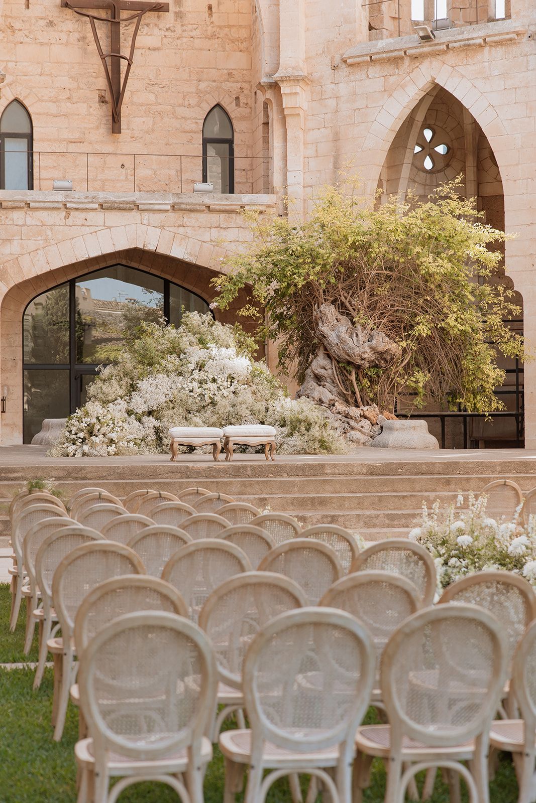 Elegant outdoor wedding ceremony setup with cane chairs and white floral arrangements at a stone venue.