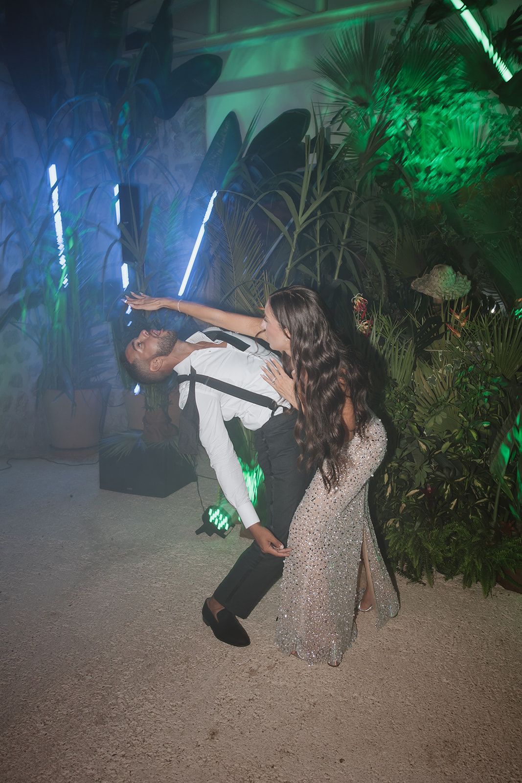 Couple dancing dramatically under green and blue lights with tropical plants in the background.