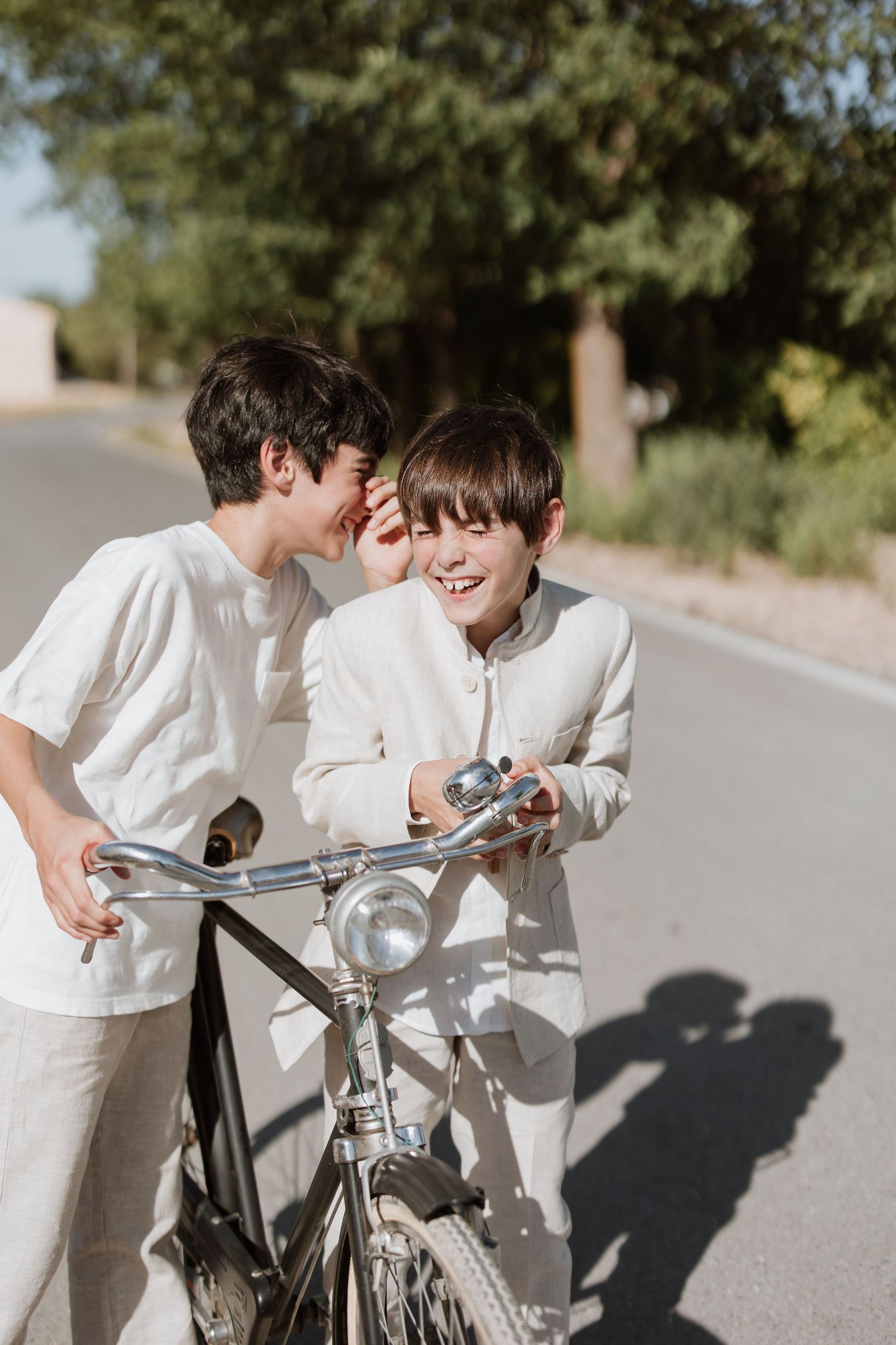 Two people in white outfits share a lighthearted moment with a bicycle on a sunny road.