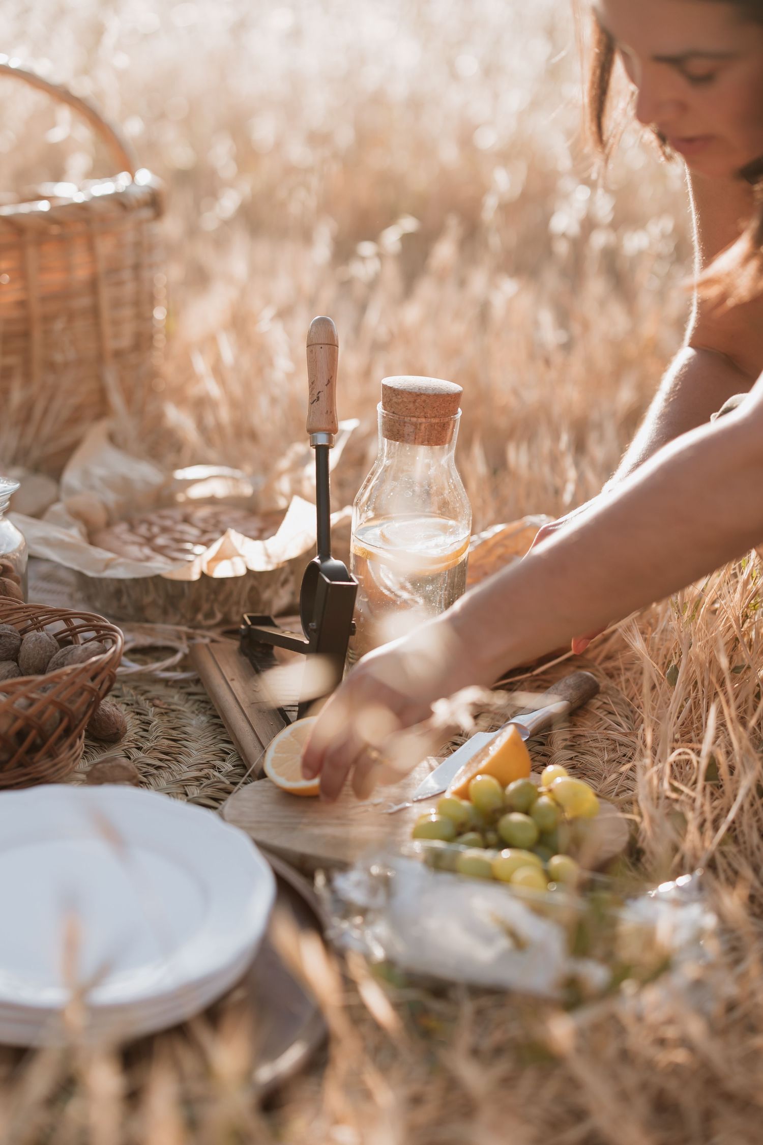 A rustic picnic scene in a wheat field with wine, grapes, and plates being set up on a blanket in warm sunlight.