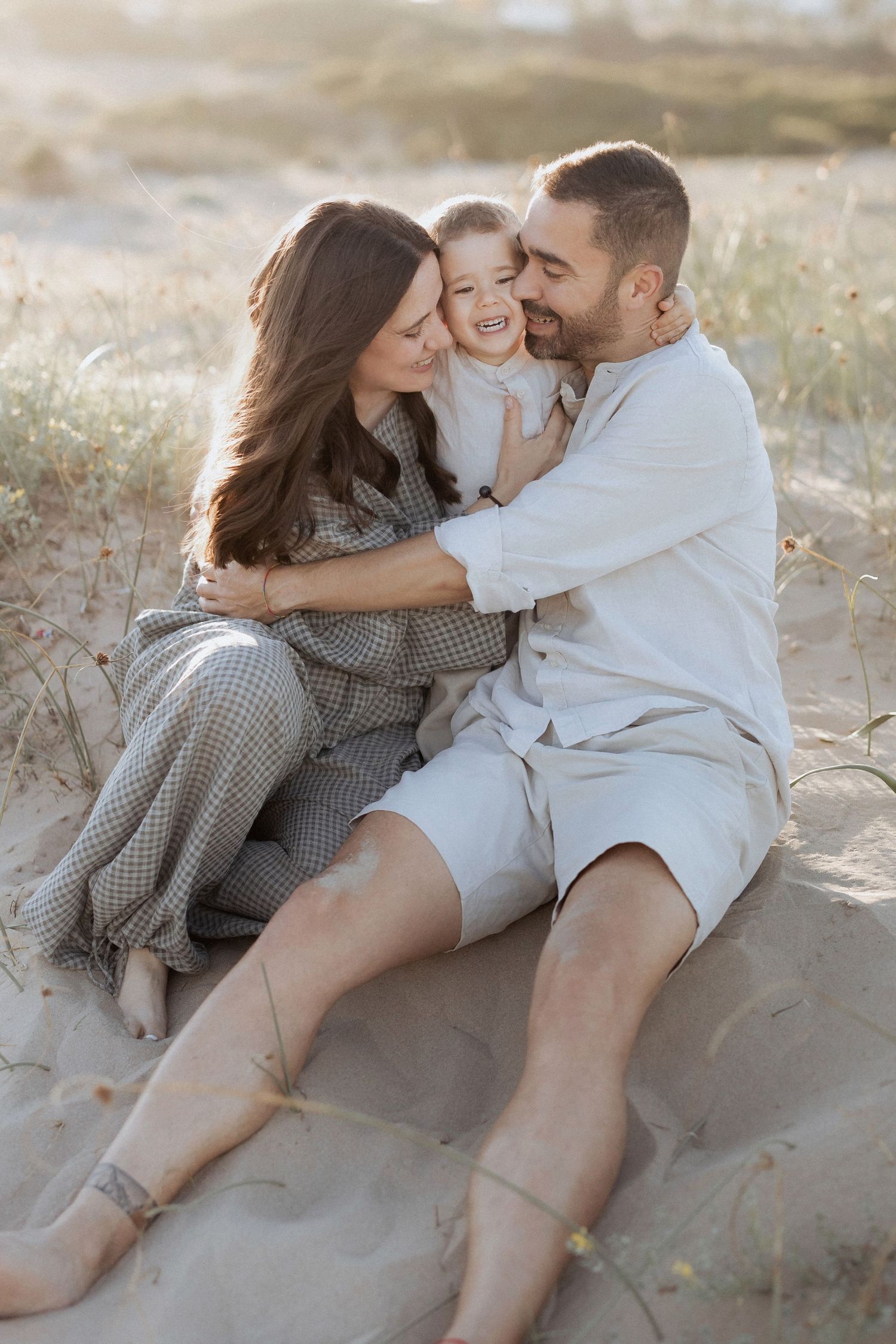 Happy family of three sitting on sandy beach dunes, parents embracing laughing toddler in warm golden light.