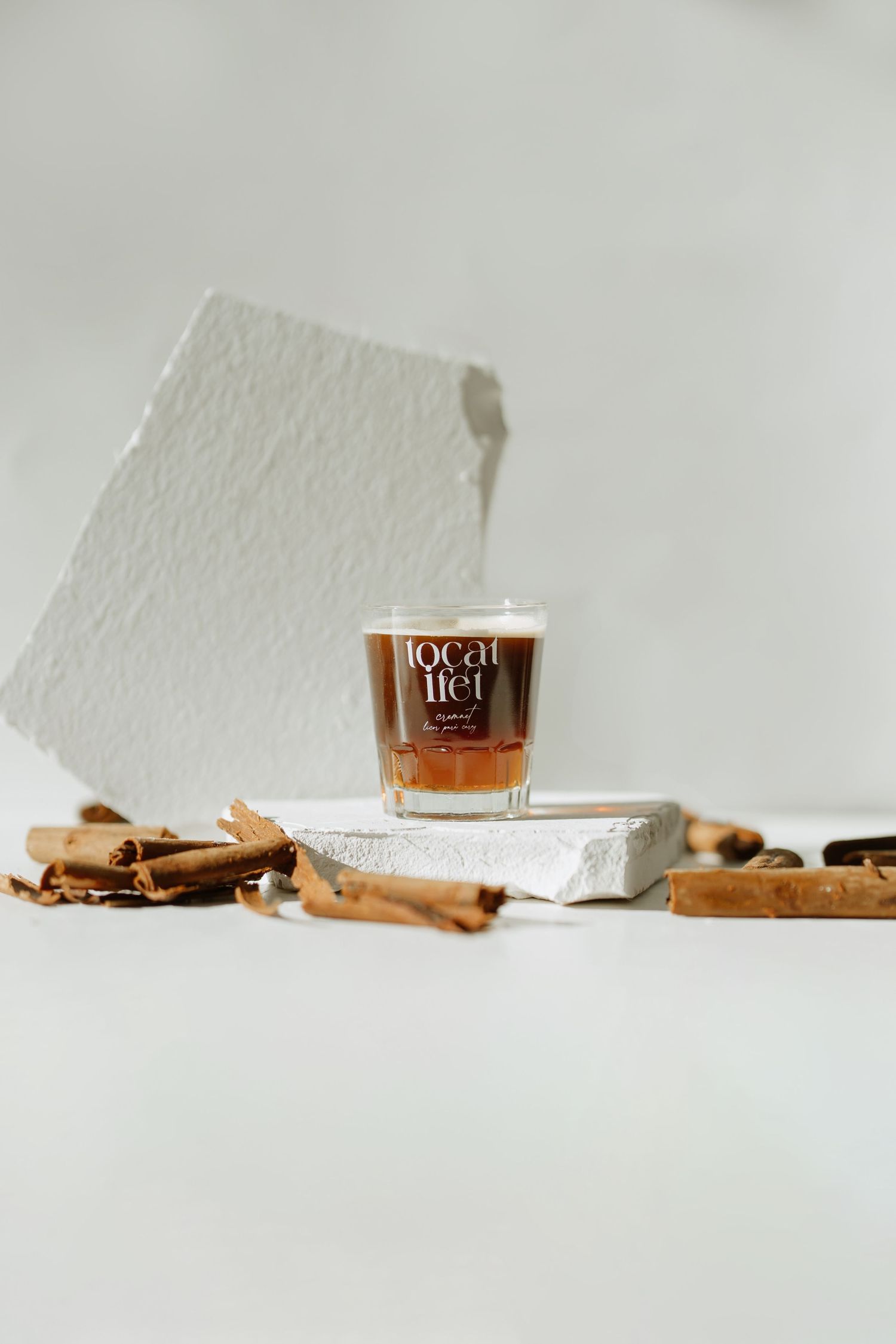 A glass of liquor with cinnamon sticks on a white pedestal against a minimalist background.