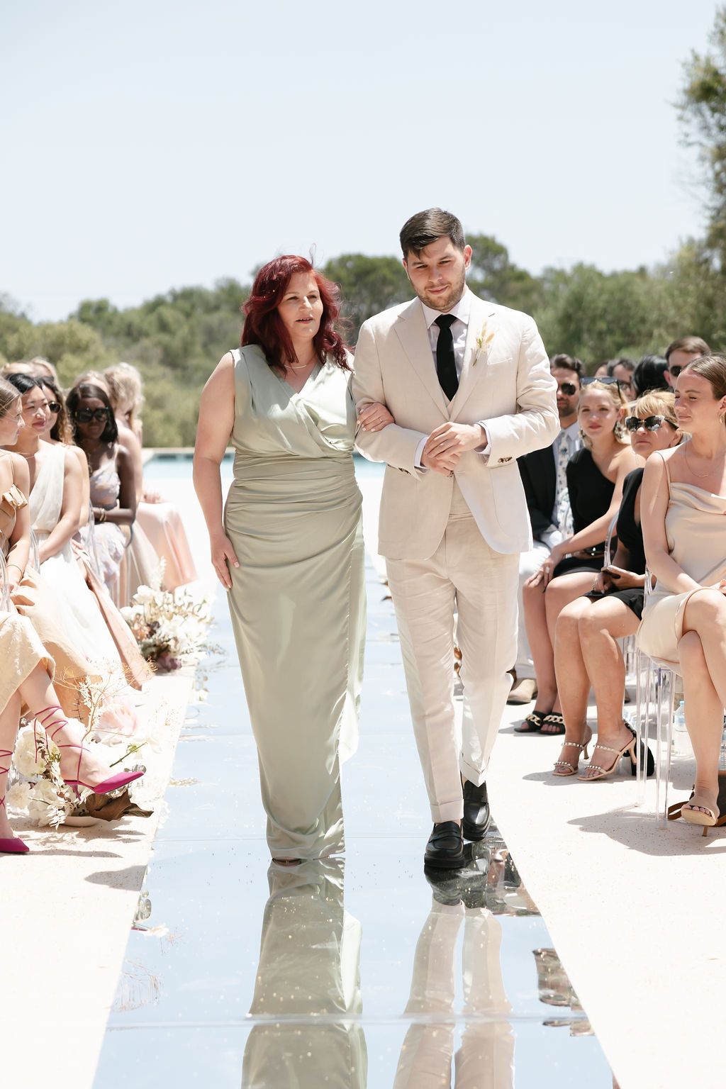 Wedding processional on mirrored aisle with attendants walking between seated guests.