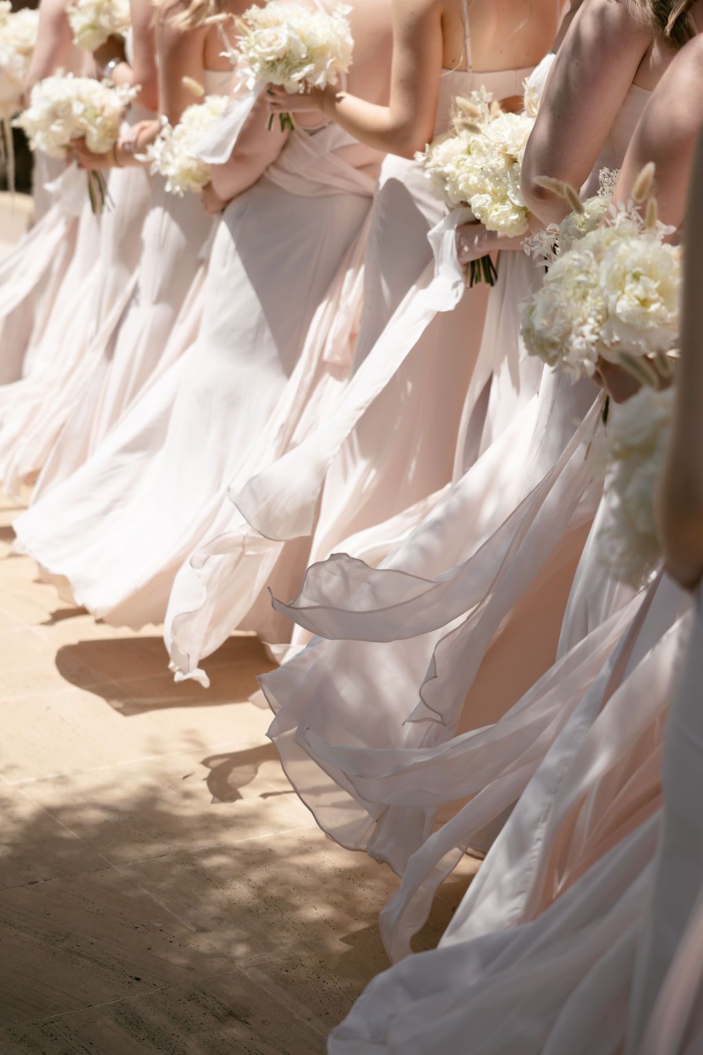 Row of bridesmaids in light pink dresses holding white bouquets at an outdoor wedding ceremony.