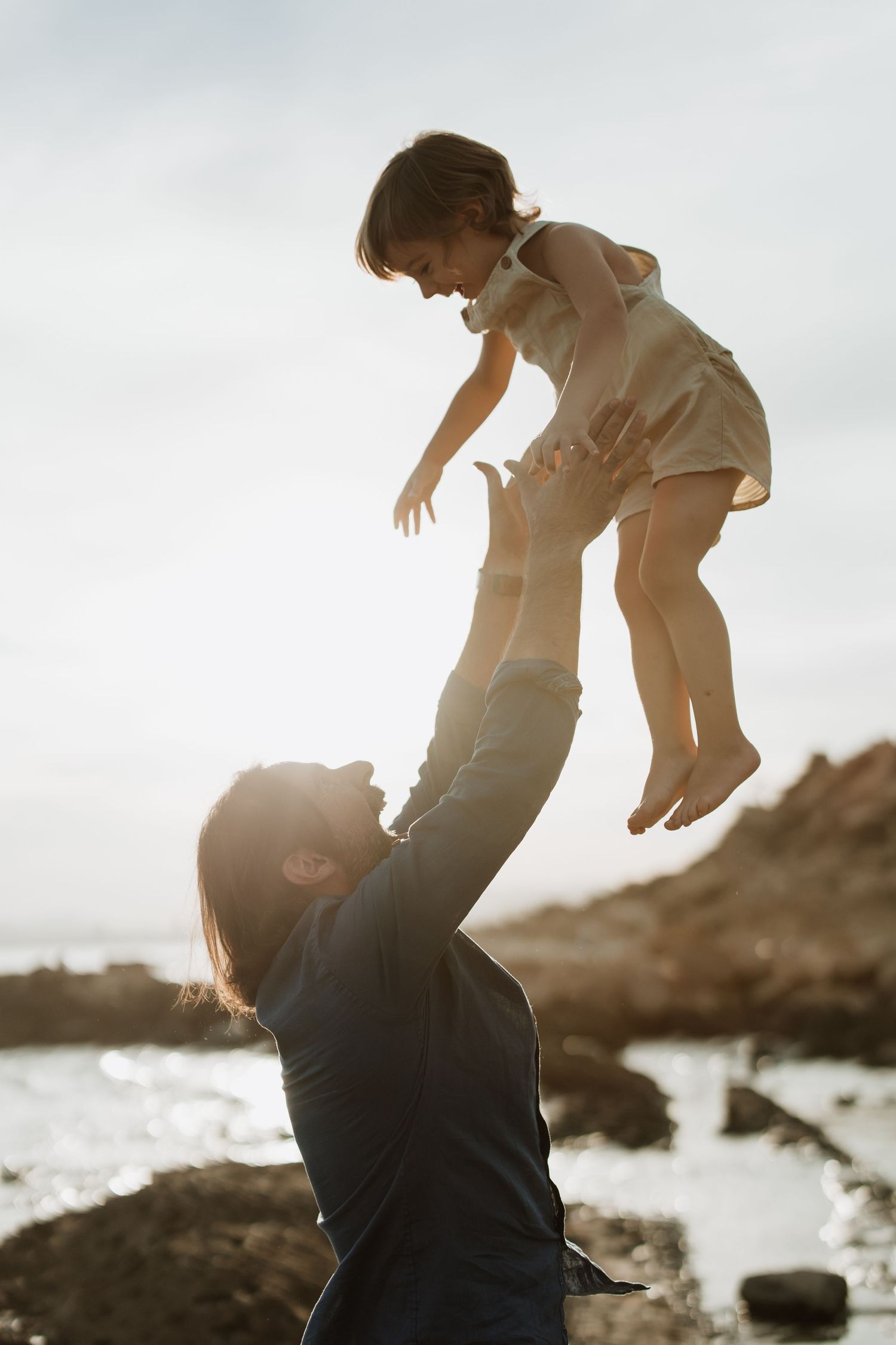 Playful moment of lifting child high in air against bright coastal background.
