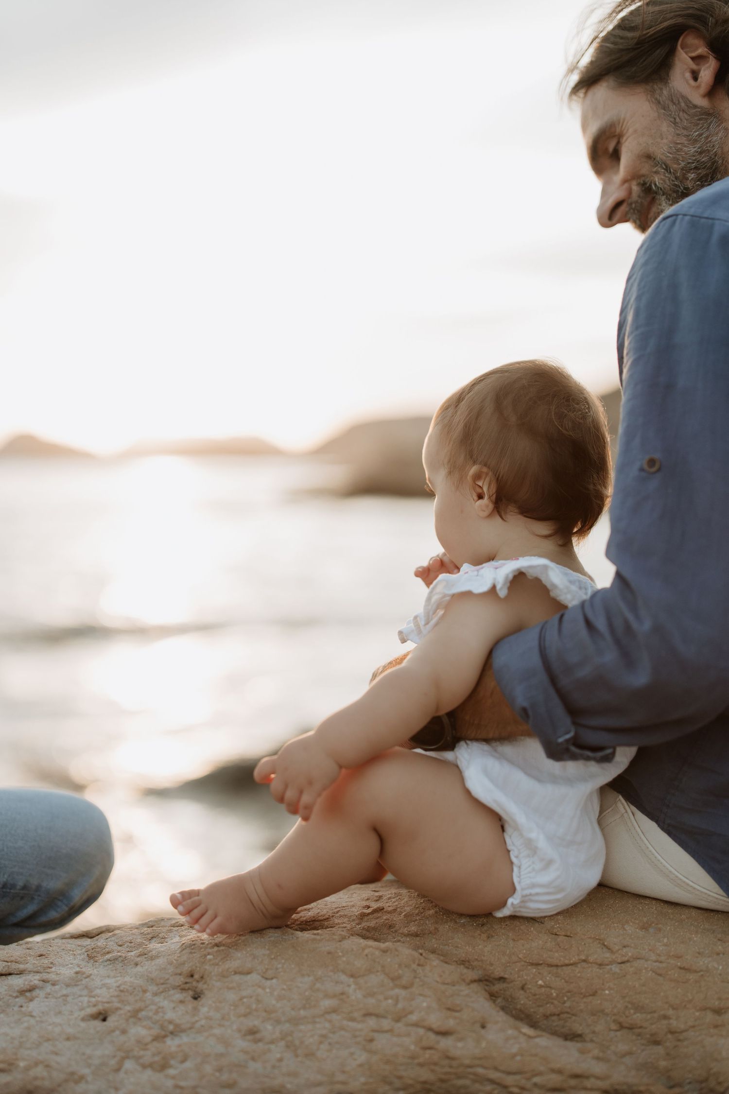 Parent and baby sharing tender moment while sitting on coastal rocks at sunset.