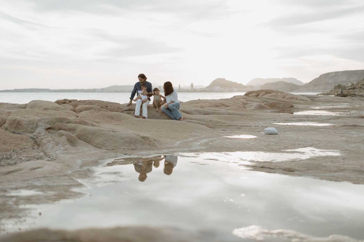 Serene coastal scene with people sitting on rocks overlooking bay and mountains.