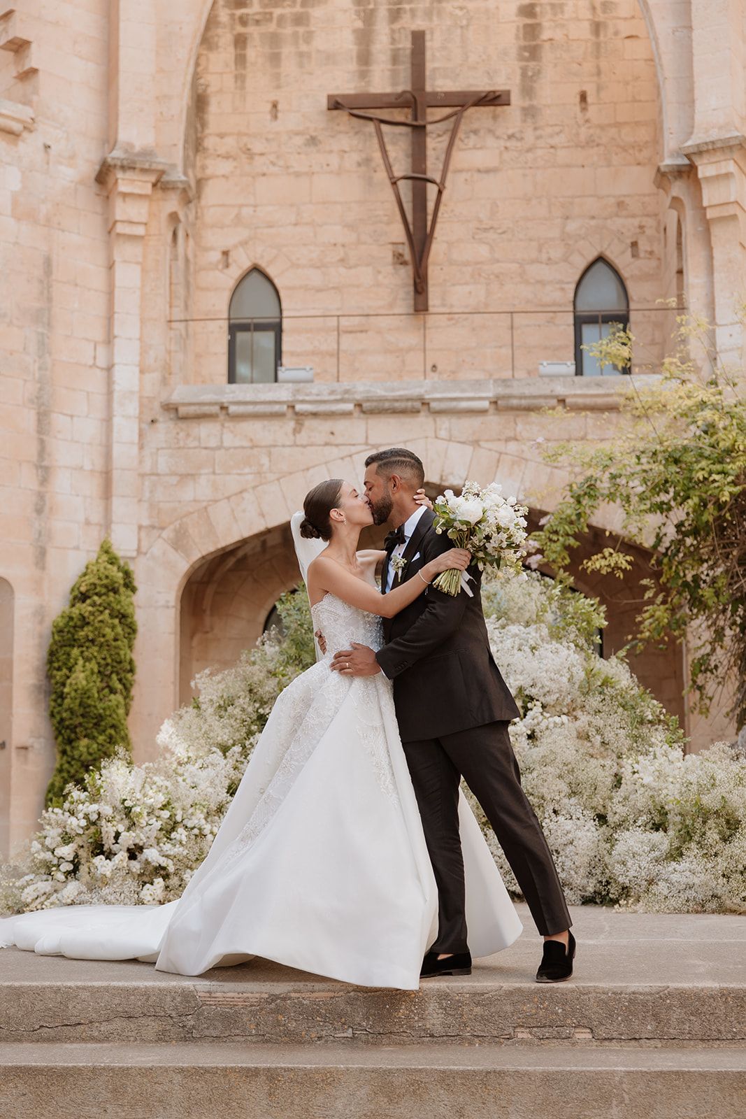 Wedding couple embracing on stone steps with white floral arrangements and gothic architecture backdrop.