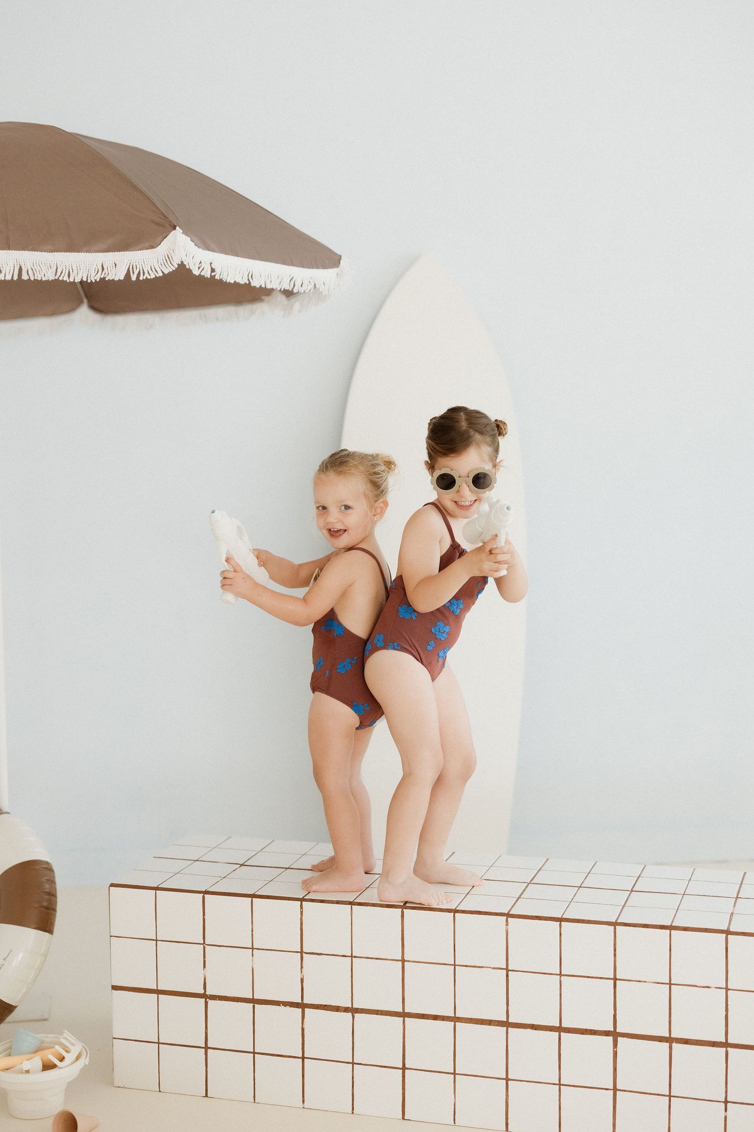 Two young girls in matching brown swimsuits stand back-to-back on a tiled ledge near a surfboard and beach umbrella.