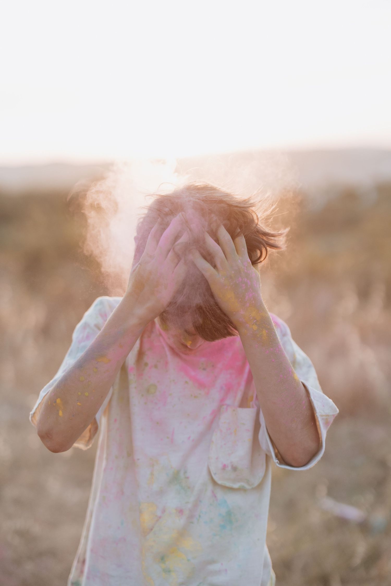Person in white shirt covered in colorful powder poses with hands covering face in sunlit golden field.