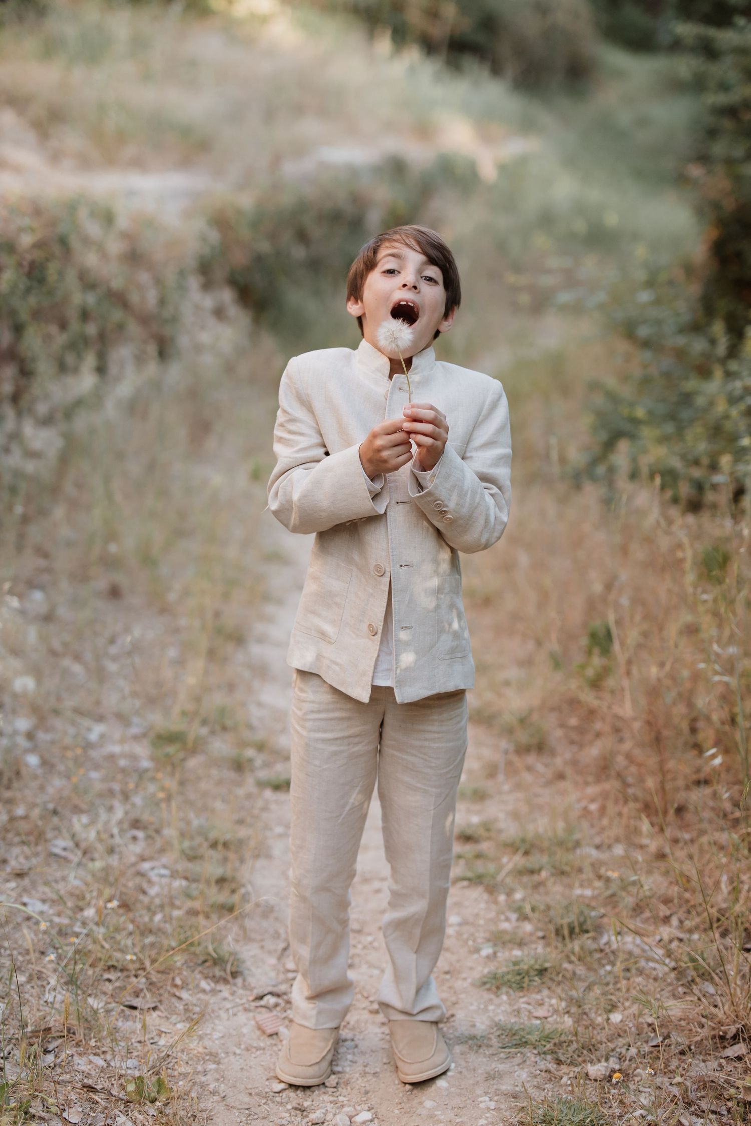 A young ring bearer in a beige suit expresses pure joy and excitement while standing outdoors on a natural pathway.