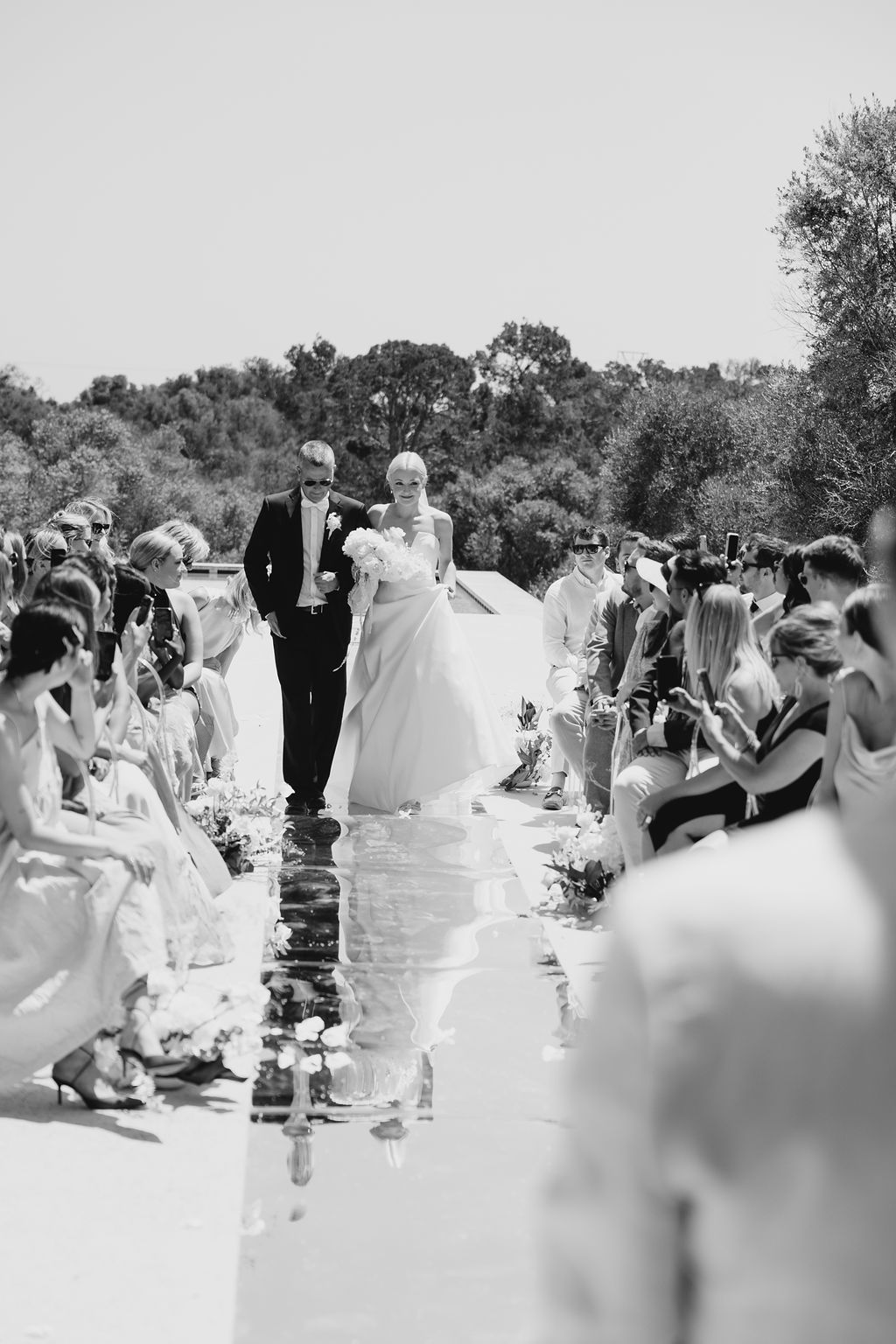 Black and white photo of wedding ceremony with guests seated along reflective aisle.