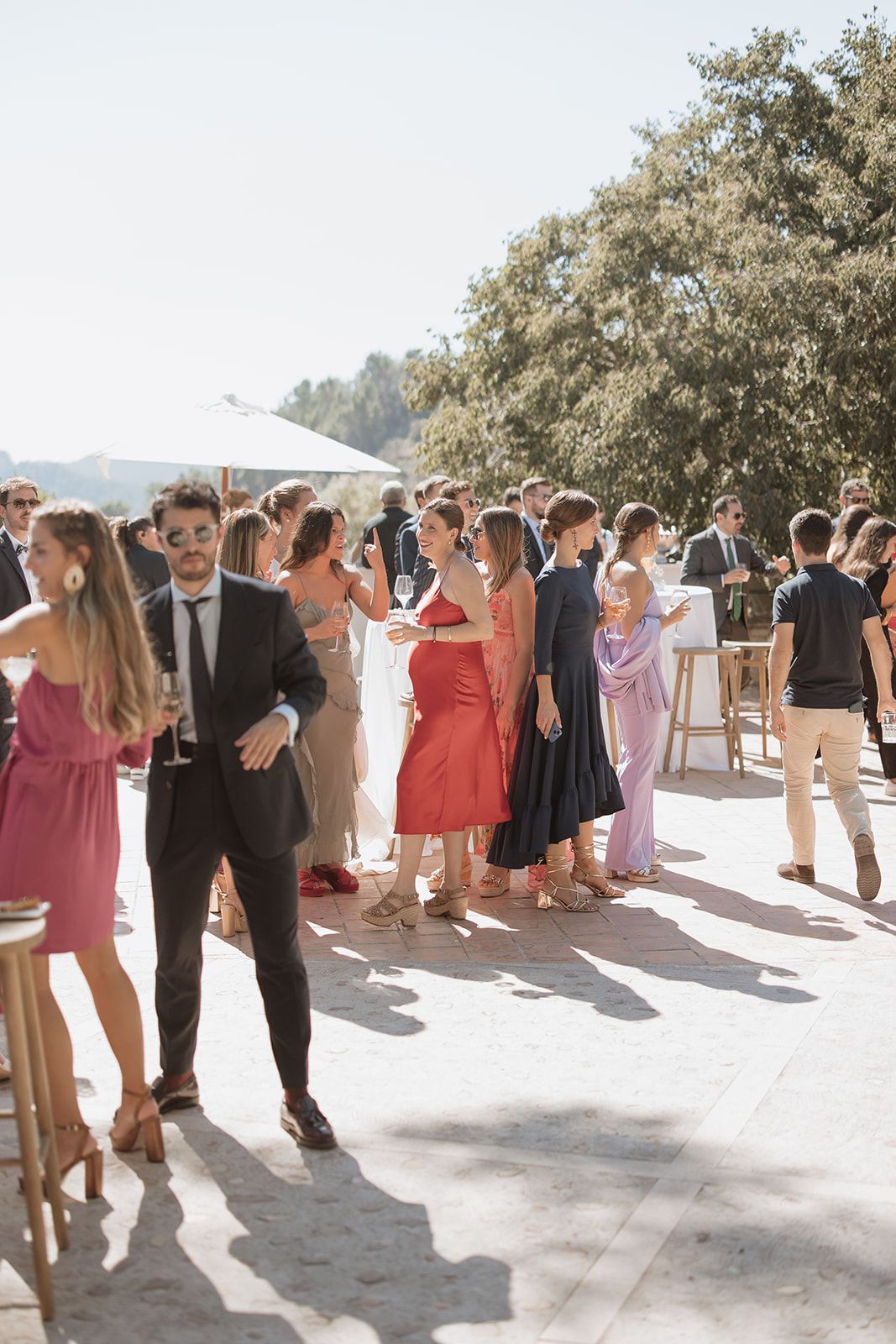 Wedding guests in colorful dresses socialize during sunny outdoor cocktail hour.