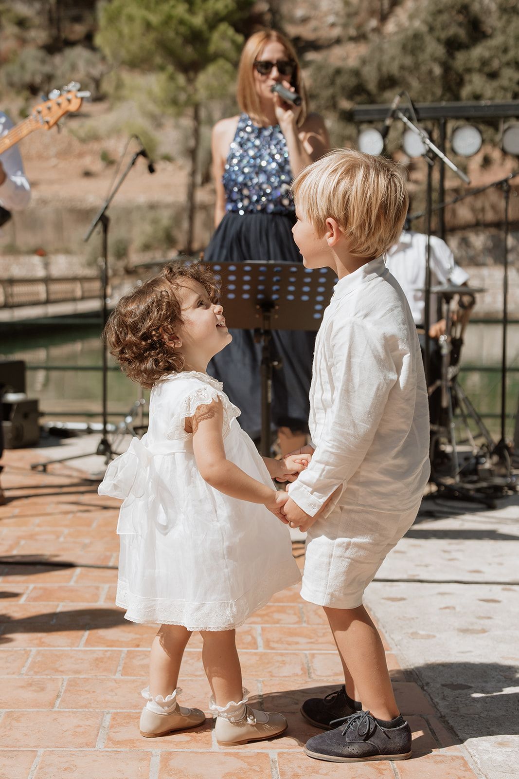 Two young children in white outfits dancing together at an outdoor event with band performing.