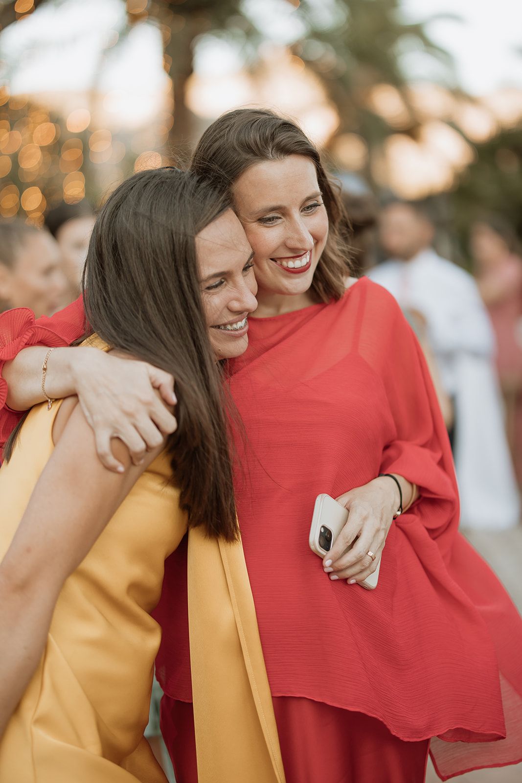 Two friends share a joyful embrace while wearing vibrant yellow and red outfits at an outdoor event.