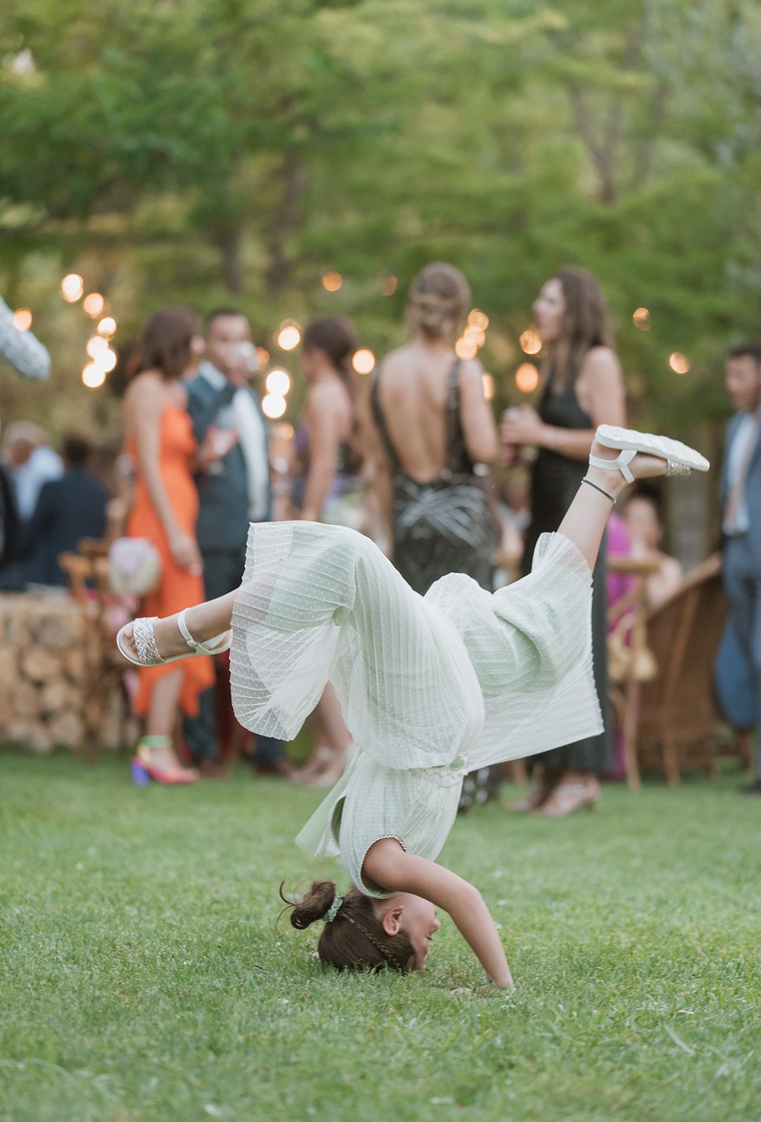 Person in white dress performing cartwheel on grass at outdoor wedding reception.