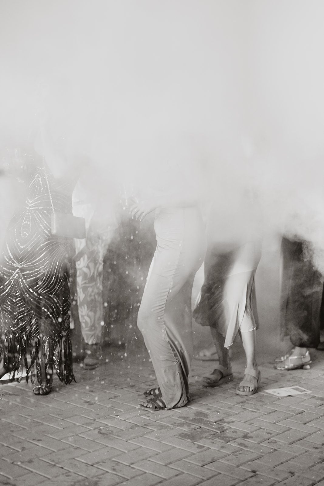 Black and white photo of dancers' legs on brick pavement through atmospheric smoke.