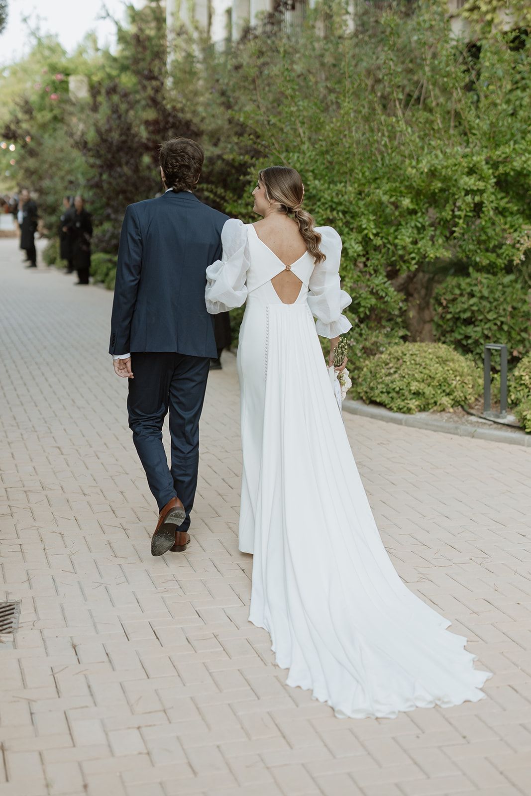 A flowing white wedding dress with open back detail as couple walks down stone path.