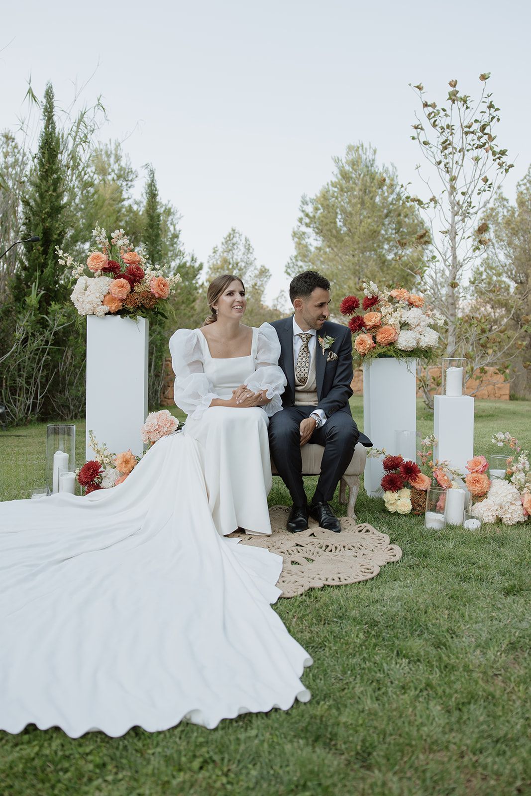 Elegant outdoor wedding scene with couple seated among white pillars decorated with coral and peach flowers.