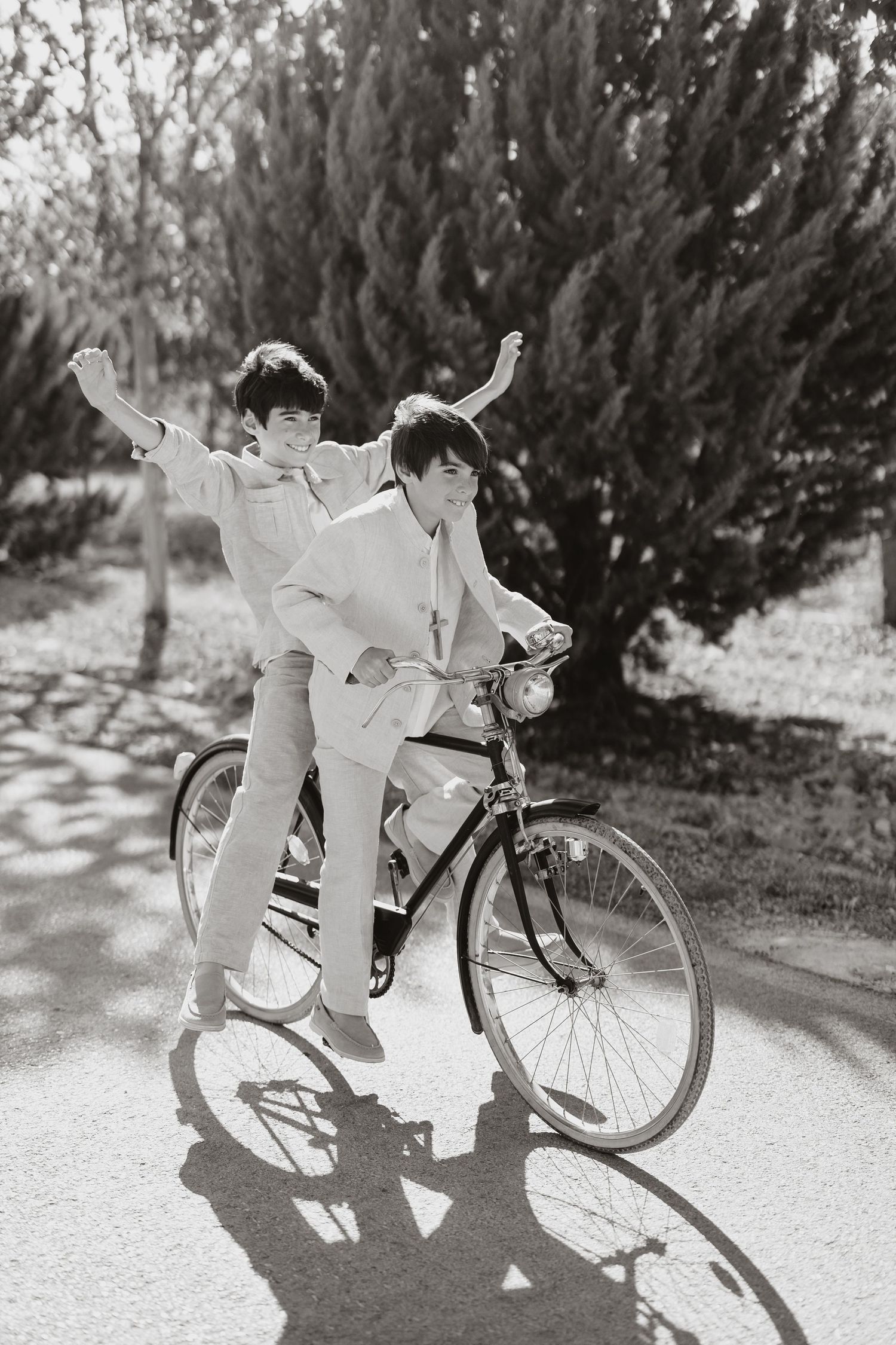 Vintage black and white photo of two people on a bicycle playing and posing on a sunny path near evergreen trees.