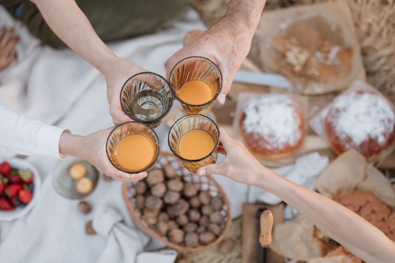 Hands toasting with orange-colored drinks in glasses over a picnic blanket with chocolate balls and baked goods.
