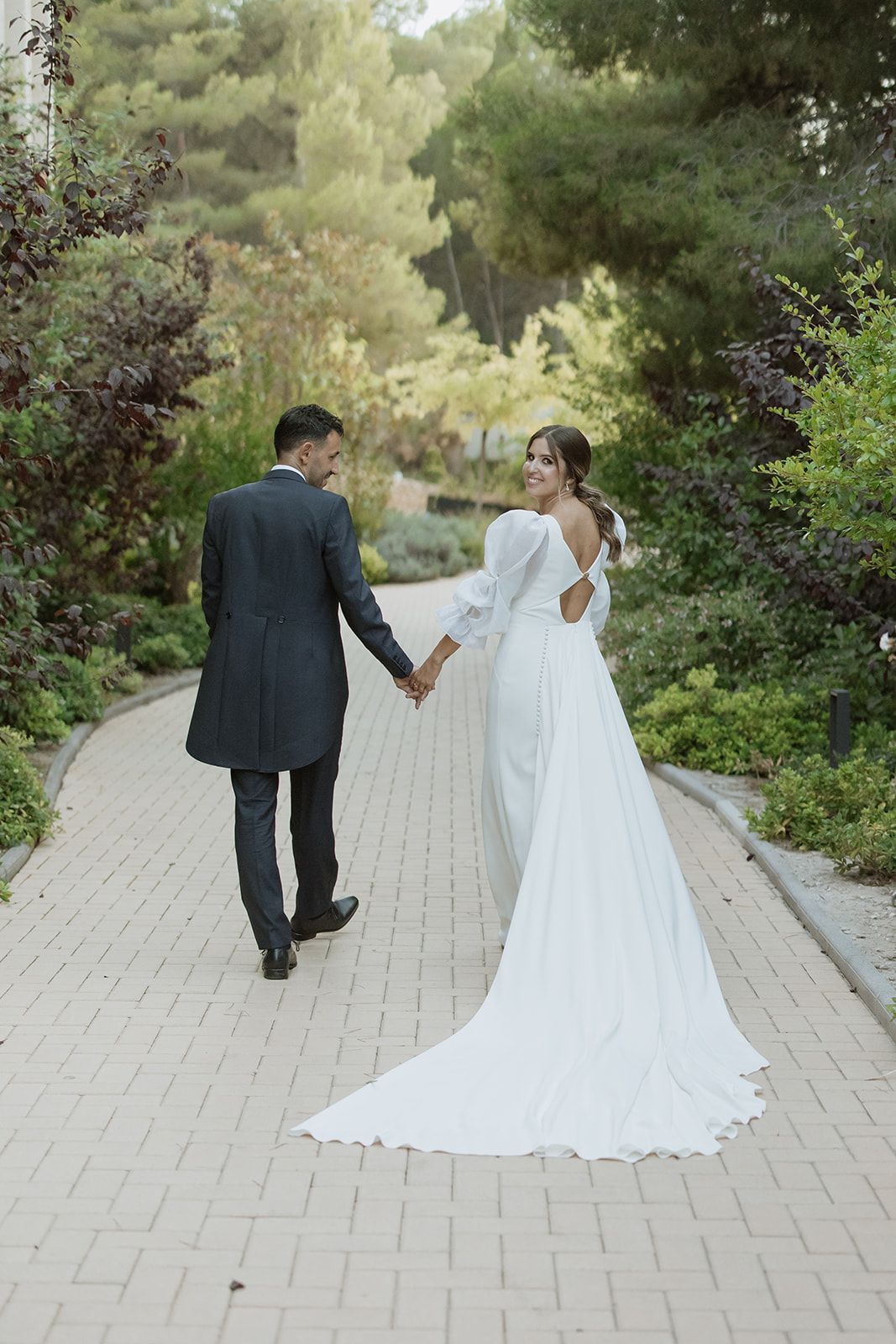 Elegant couple walks hand in hand down garden path showing bride's flowing white dress train.