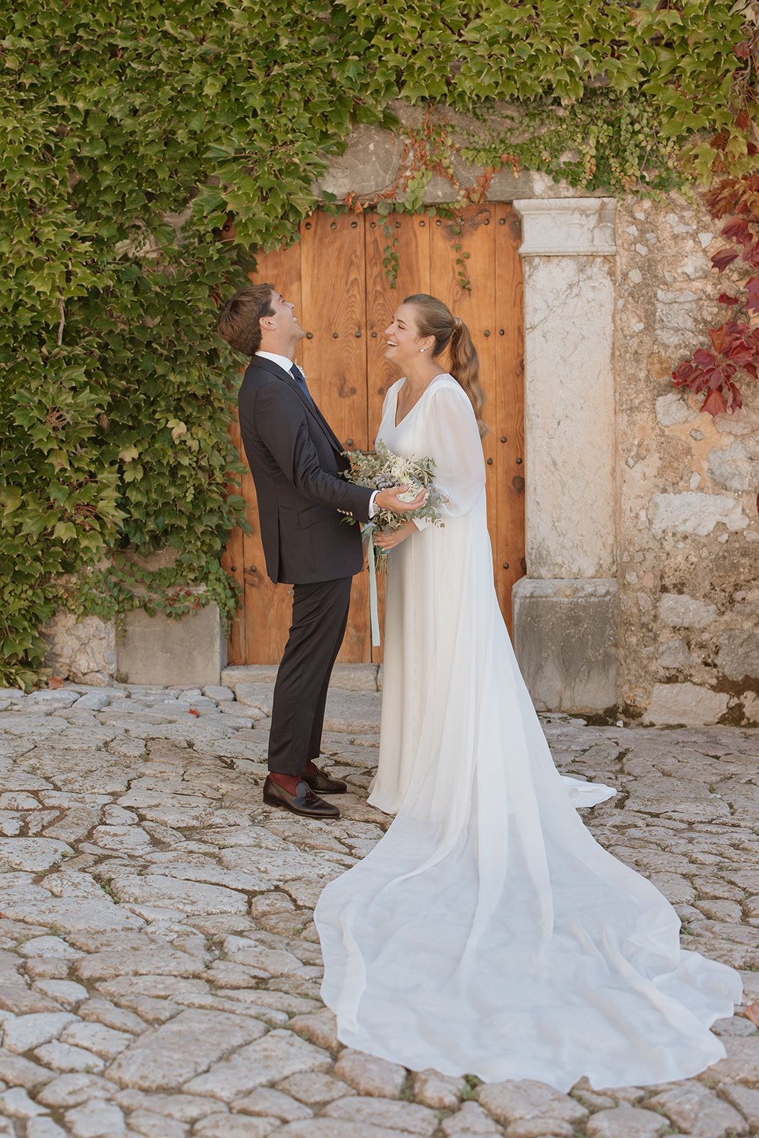 A couple in wedding attire stands before a rustic wooden door framed by climbing vines.