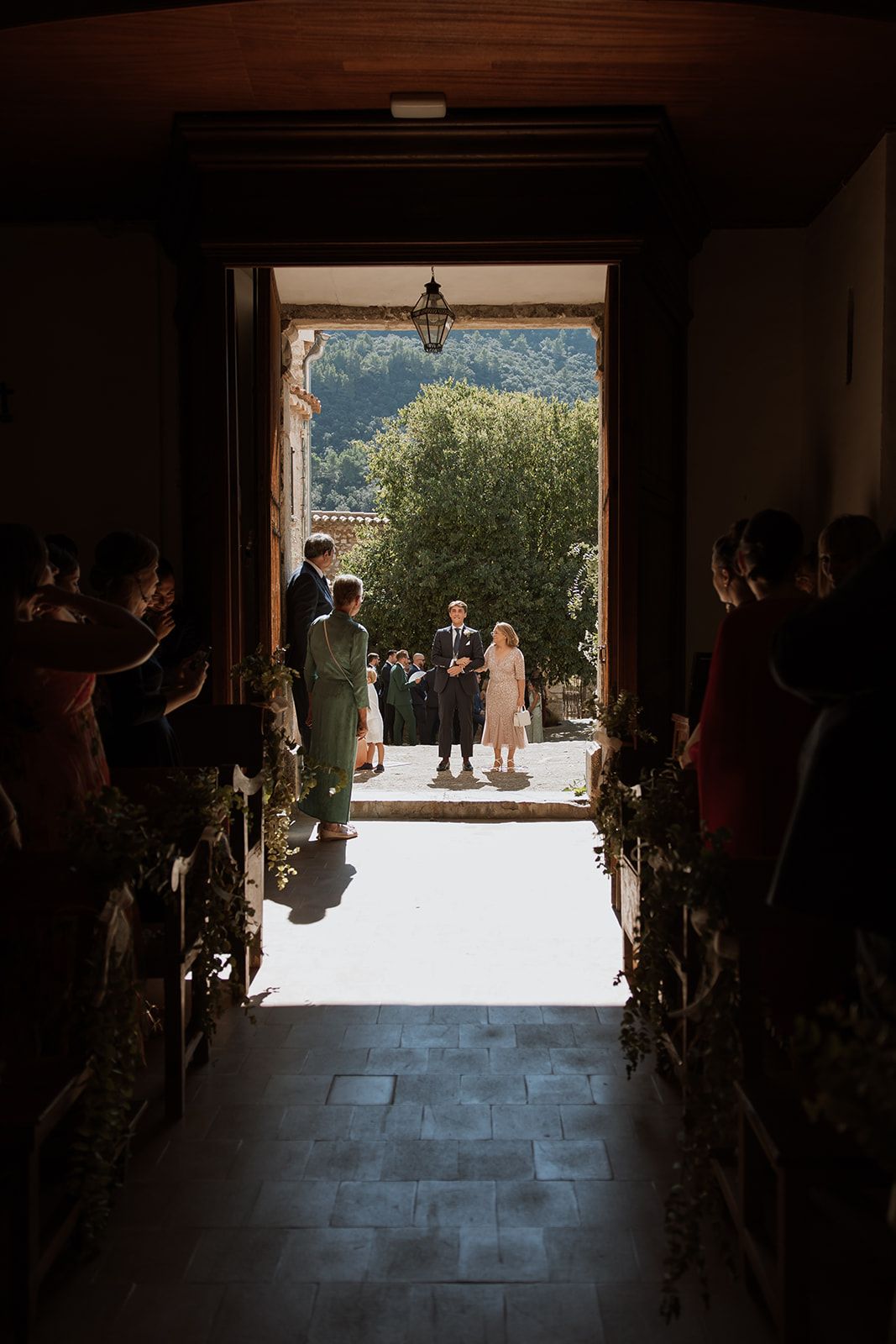 Guests watch a wedding ceremony from wooden pews as sunlight streams through church doorway.