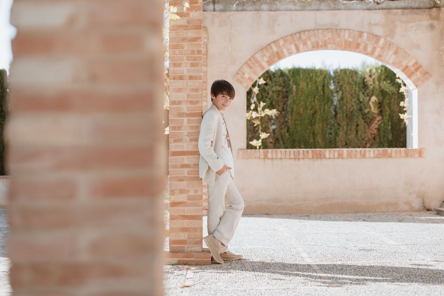 A couple stands together in a romantic pose along an elegant brick archway with garden scenery in the background.