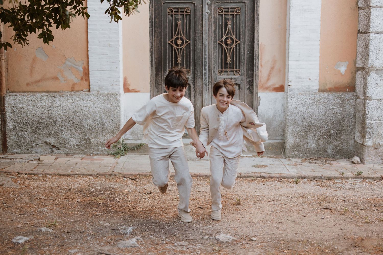 Two children in white clothing run playfully together in front of an old wooden door and textured wall.