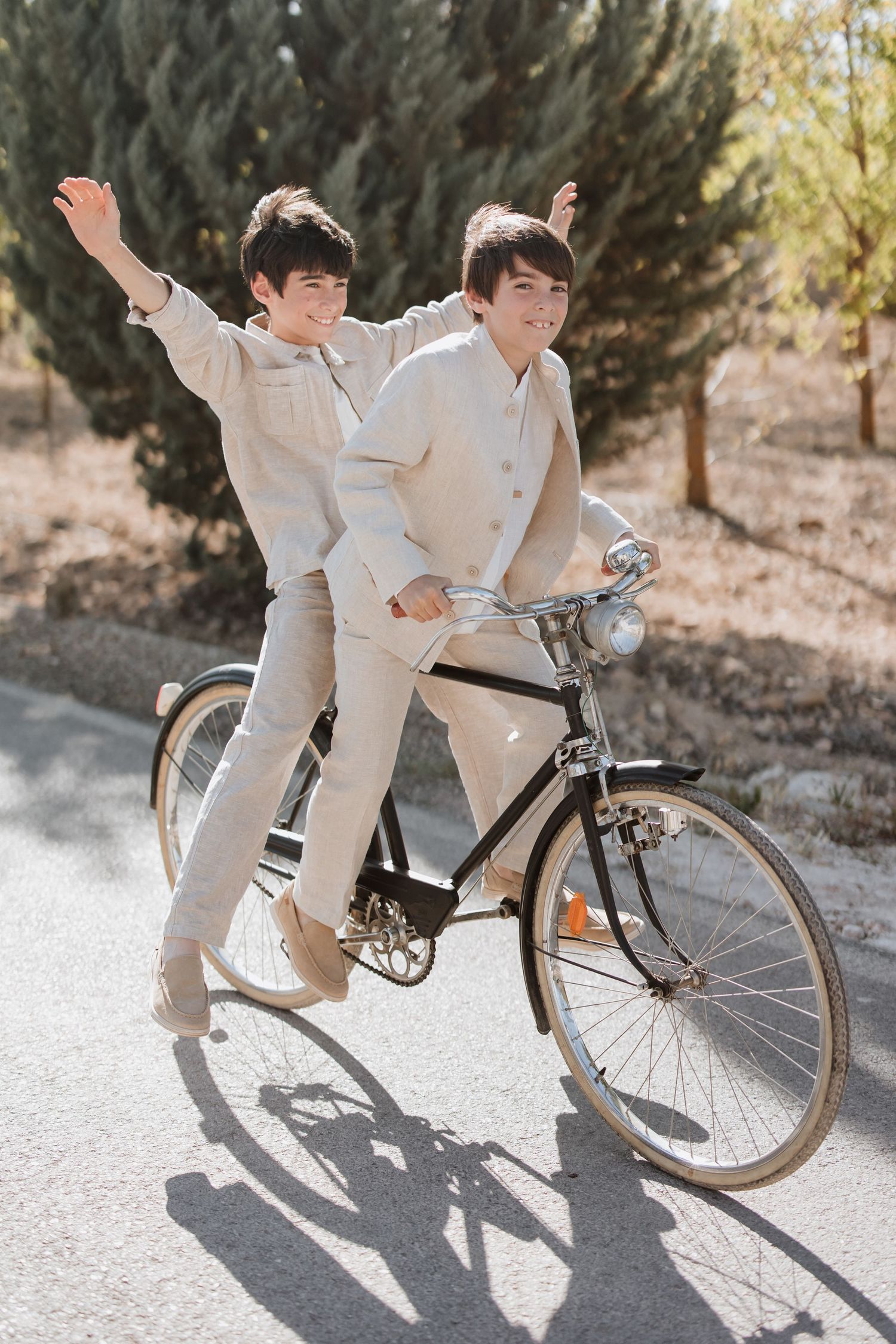 Two people in matching beige outfits playfully riding a vintage black bicycle together on a sunny day.