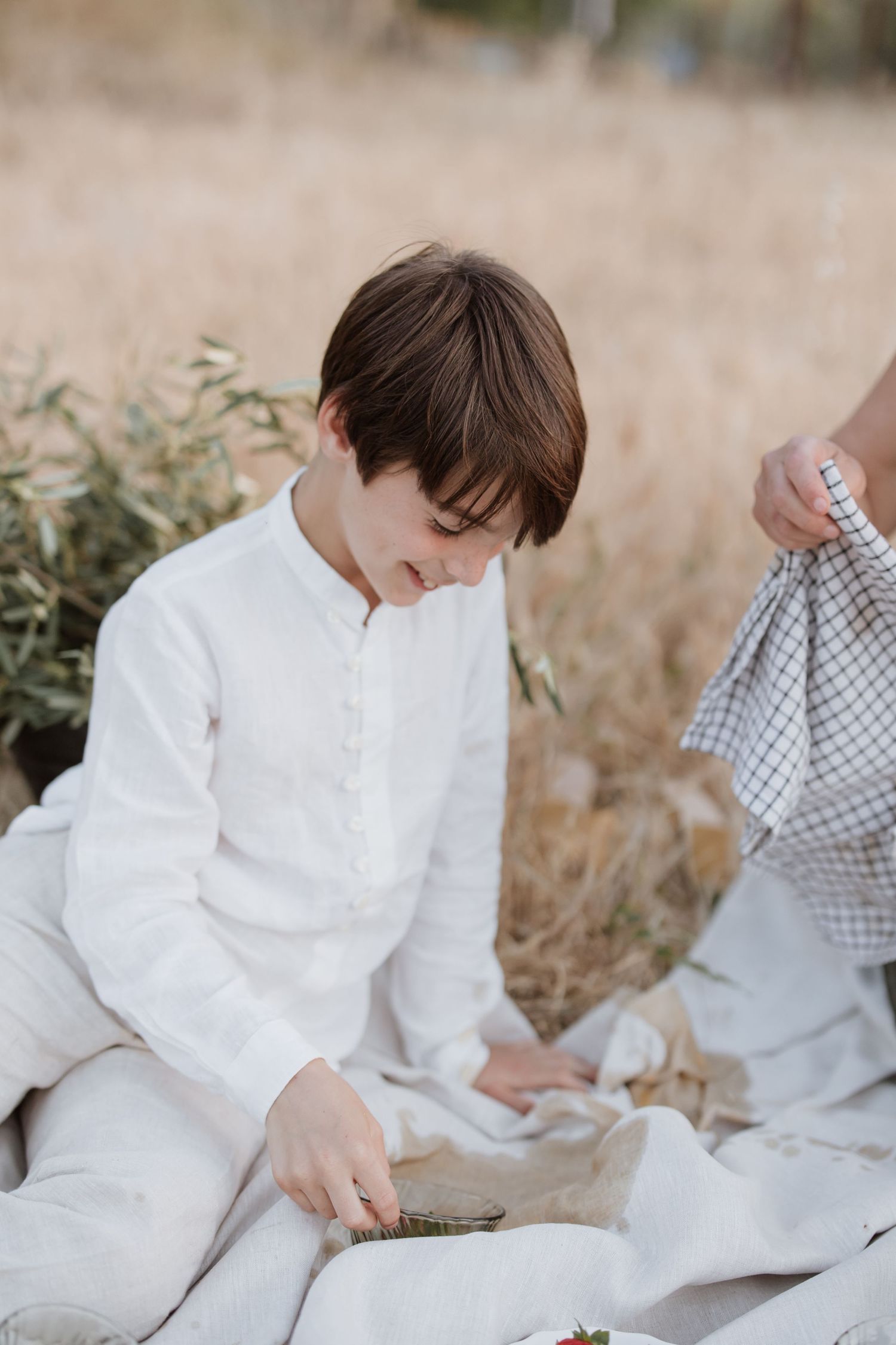 A young figure in a white linen ensemble sits thoughtfully in a natural outdoor setting with dried grass and foliage.