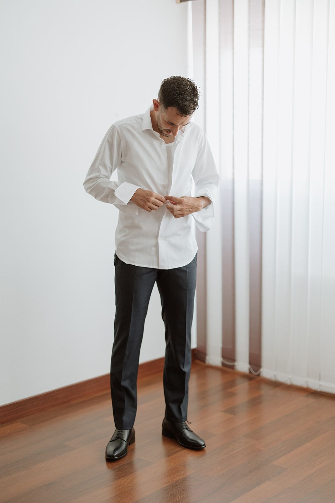 A person in white shirt and dark pants buttons their sleeves while standing in a bright room.