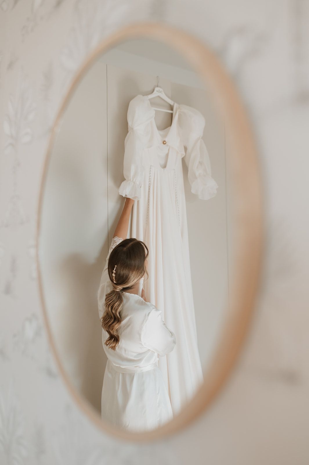 White wedding dress hanging in oval mirror reflection with someone looking up at it.