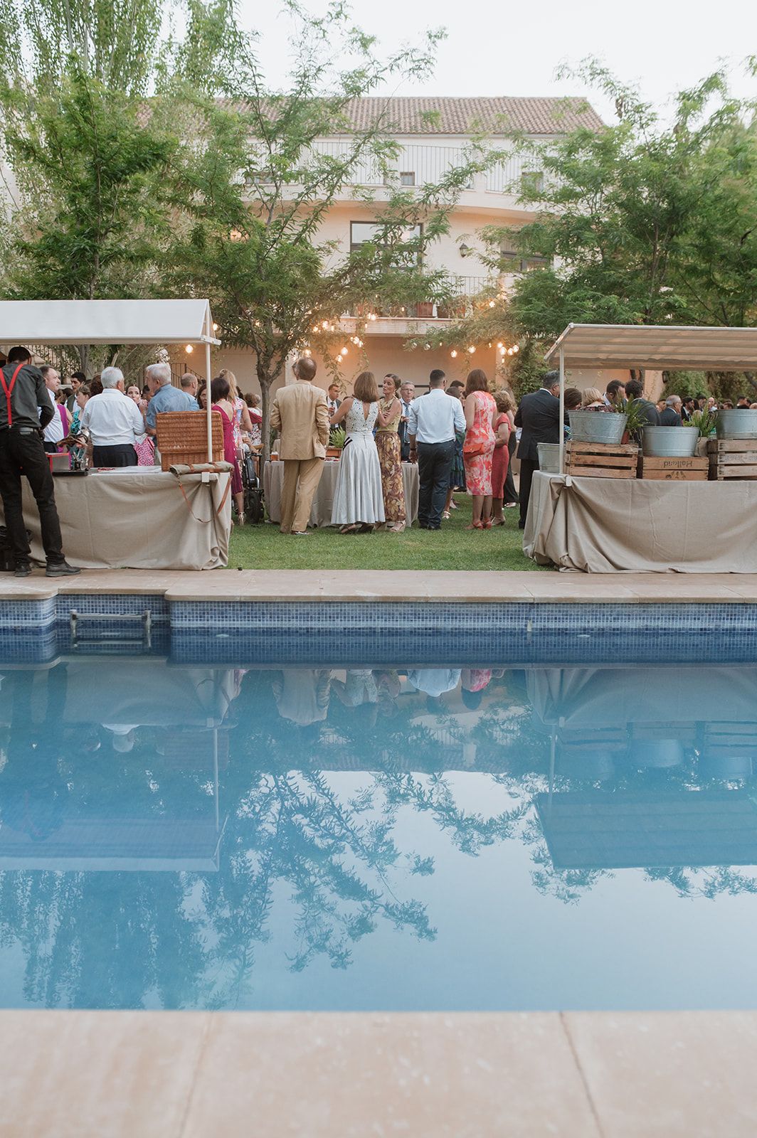 Wedding guests gather around a poolside reception area with tables and decorations at sunset.