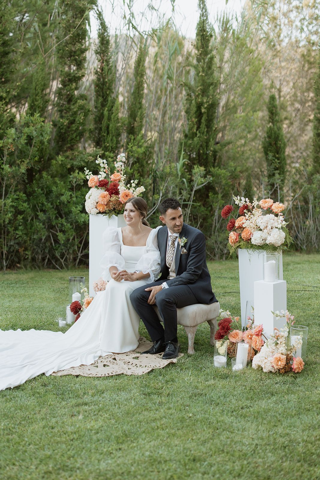 Newlyweds seated on elegant chair surrounded by floral arrangements at outdoor garden wedding.