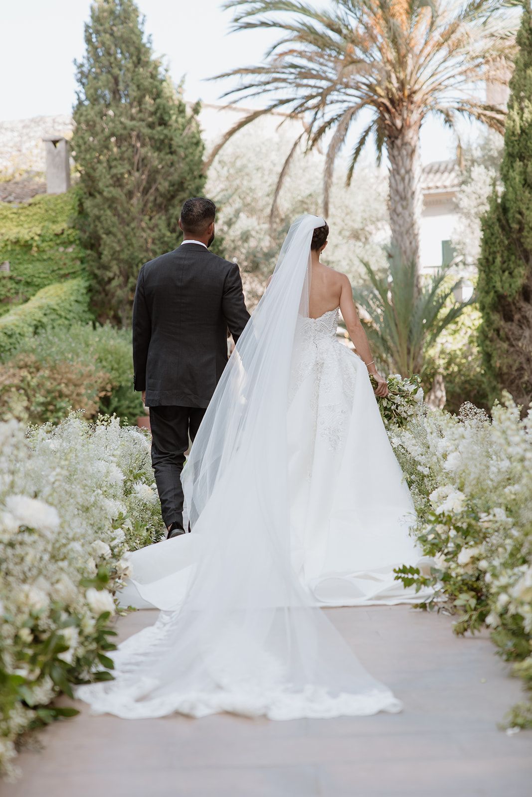 A romantic wedding moment captured in a garden path lined with palm trees and white floral arrangements.