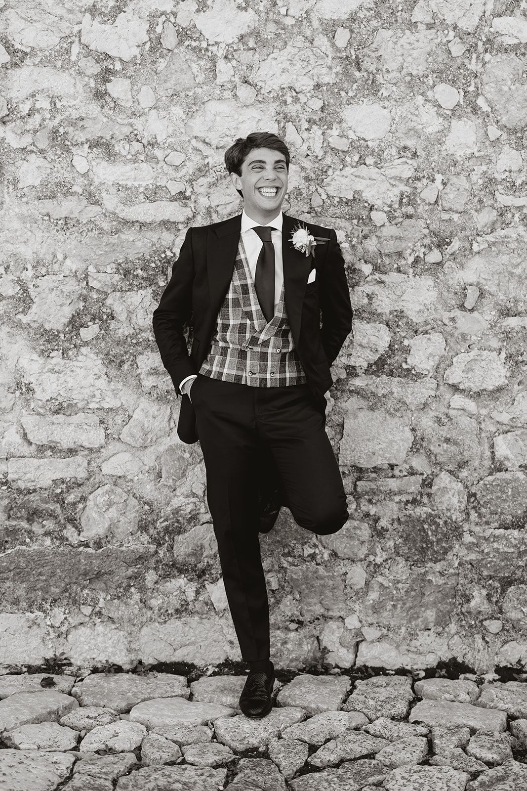 Groom in formal suit and plaid vest poses against rustic stone wall.