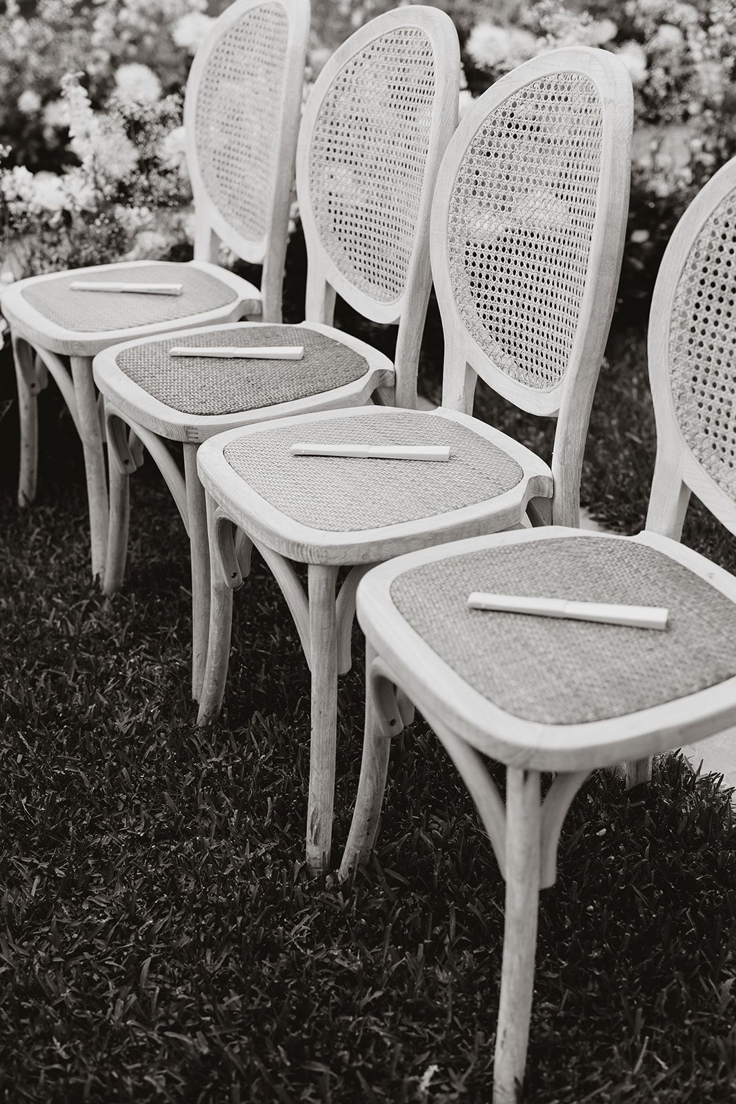 White cane-back chairs with place cards arranged in a row on grass for an outdoor event.