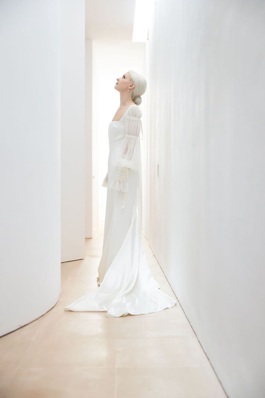 An ethereal bridal portrait in a minimalist white hallway showing an elegant long-sleeved gown.