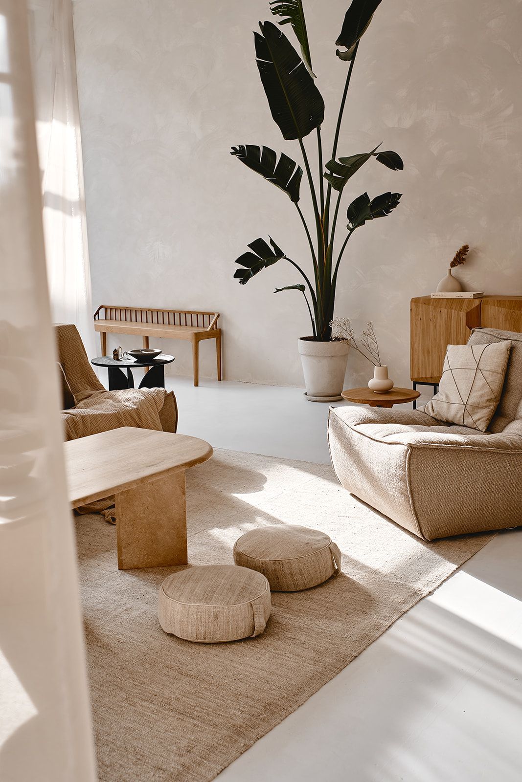 Minimalist boho living room with neutral tones, linen sofas, round coffee table, woven poufs, and a large tropical plant.