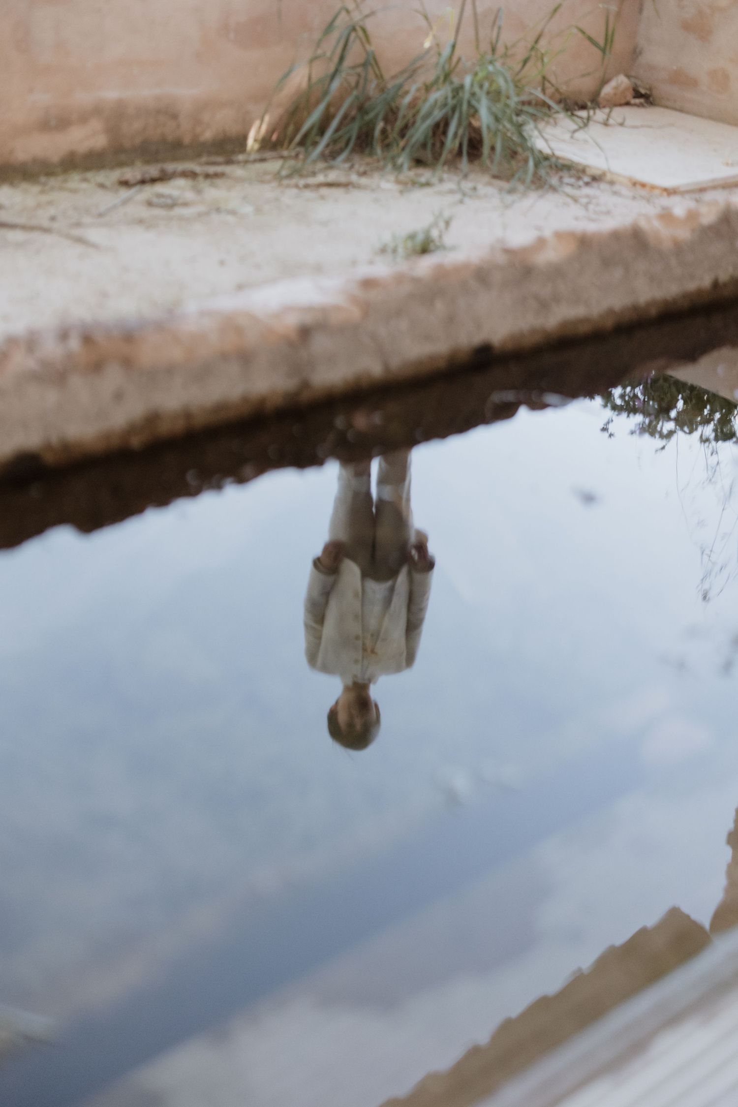 A reflection in still water shows an upside down figure against a concrete edge with grass.