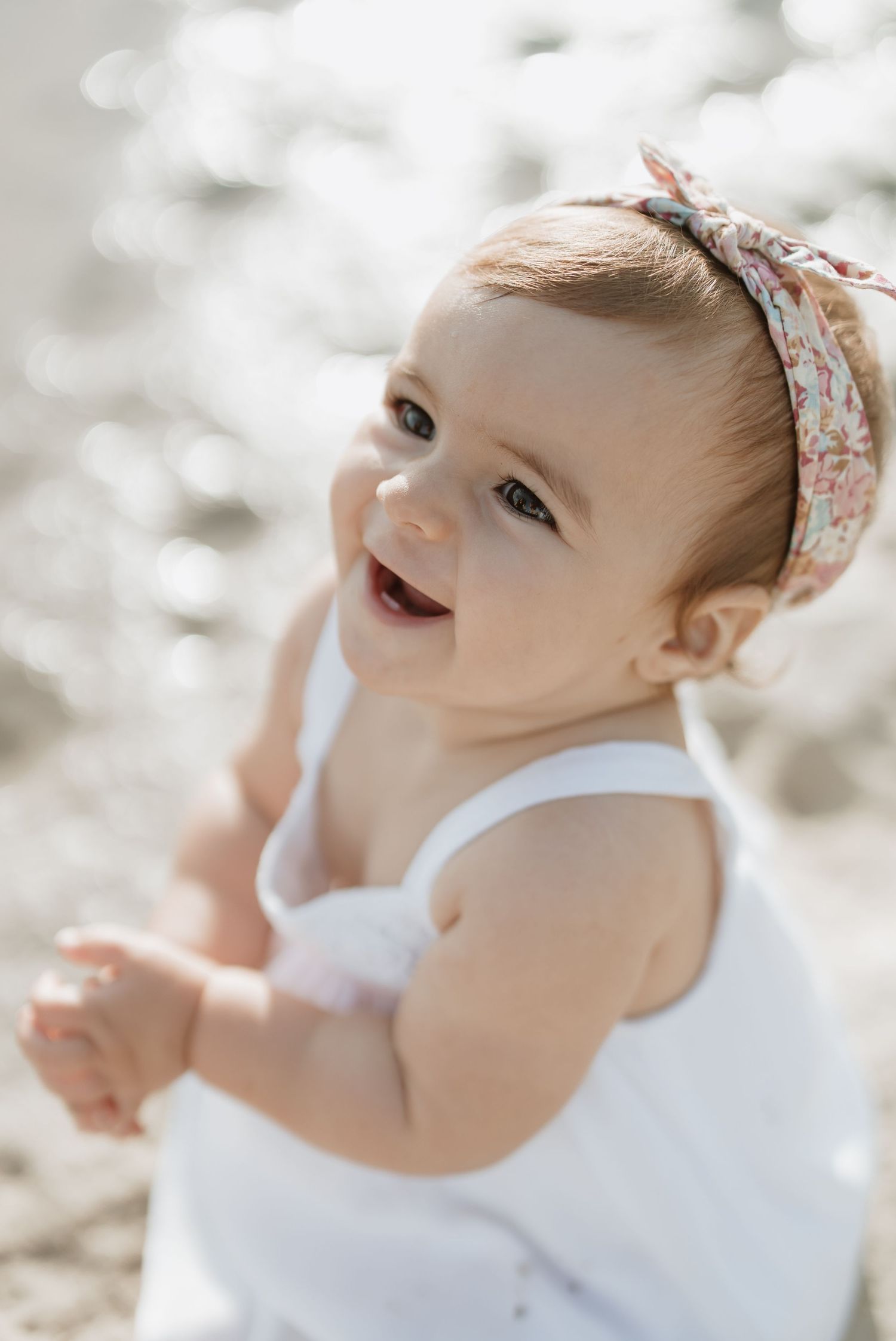 Joyful baby in white tank dress and floral headband smiles brightly outdoors.