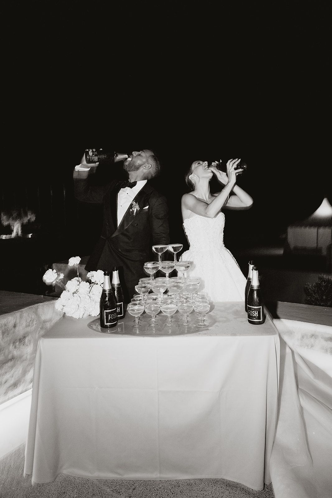 Black and white photo of newlyweds taking a celebratory drink beside their champagne tower.