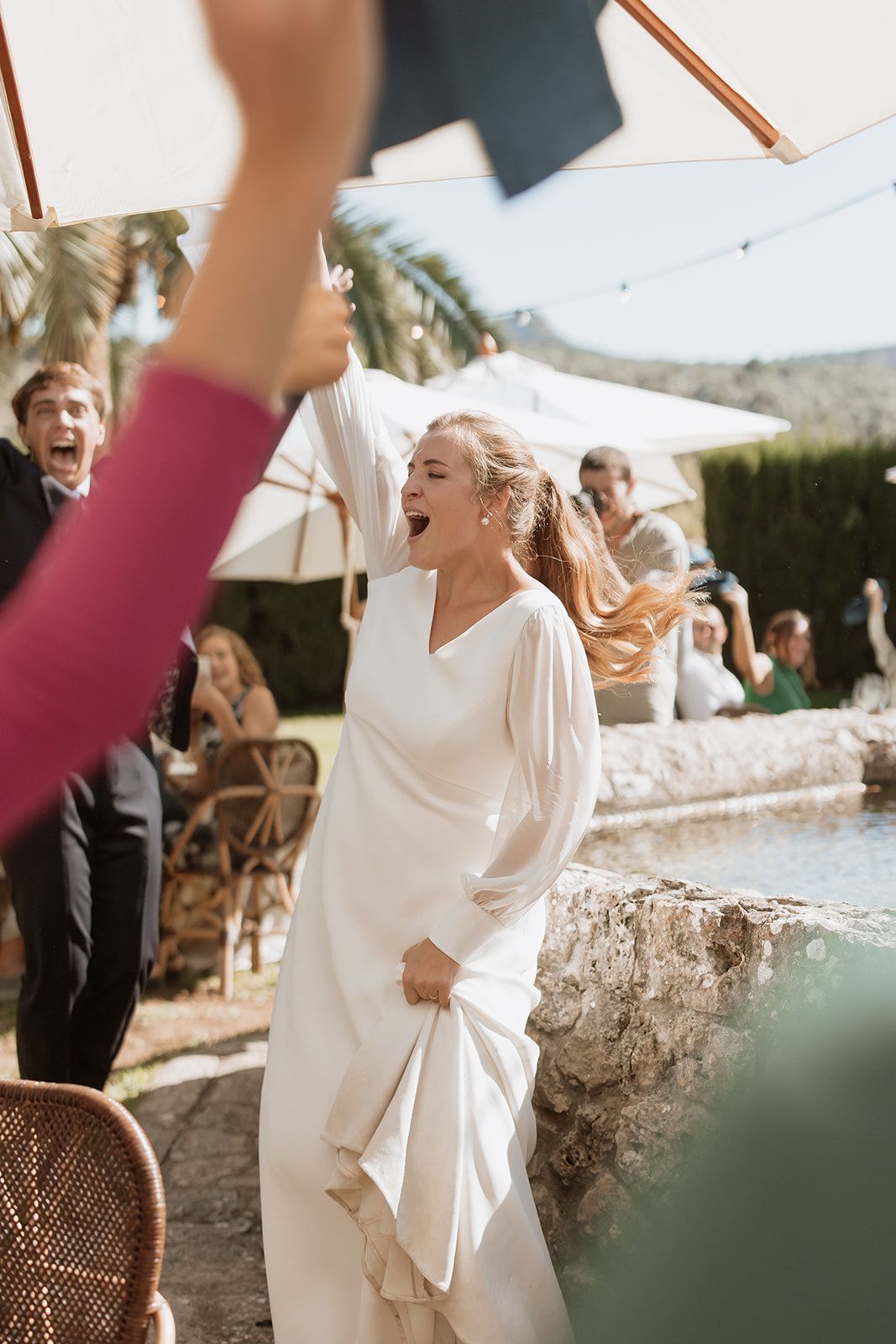 Bride in white dress celebrates joyfully during outdoor wedding reception near palm trees.