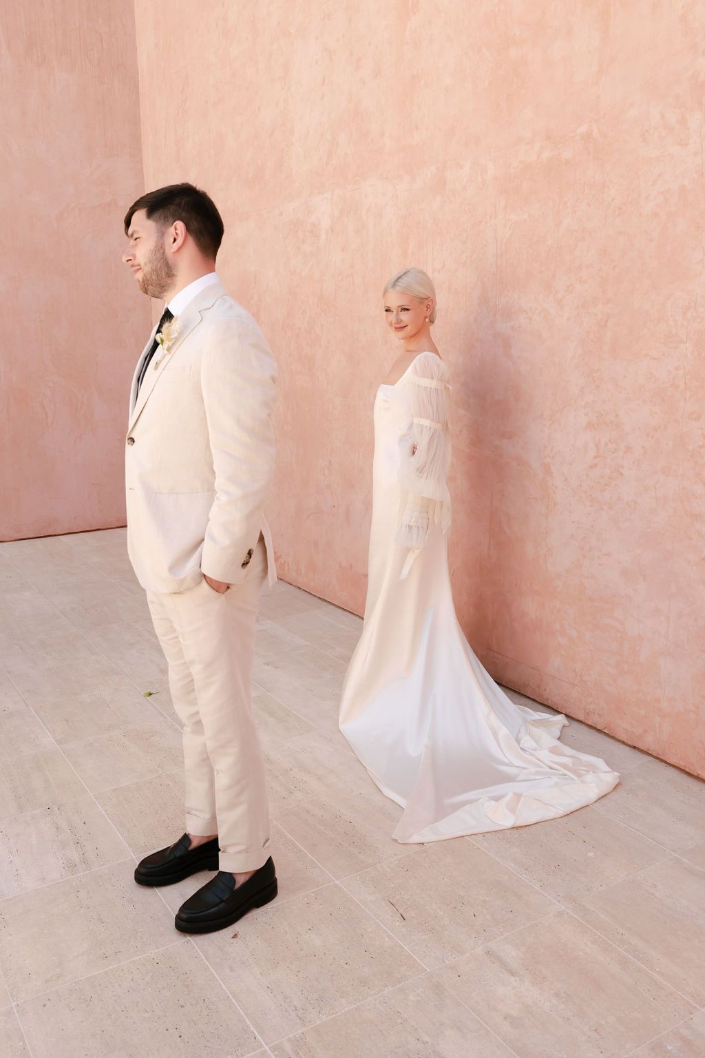 Modern wedding couple in cream-colored attire against pink wall for first look photos.