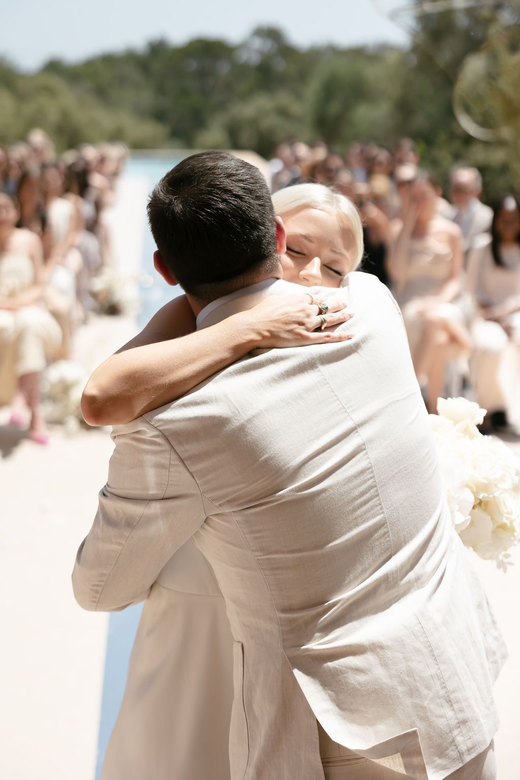 A tender embrace between two people in white attire during an outdoor wedding ceremony with guests in the background.