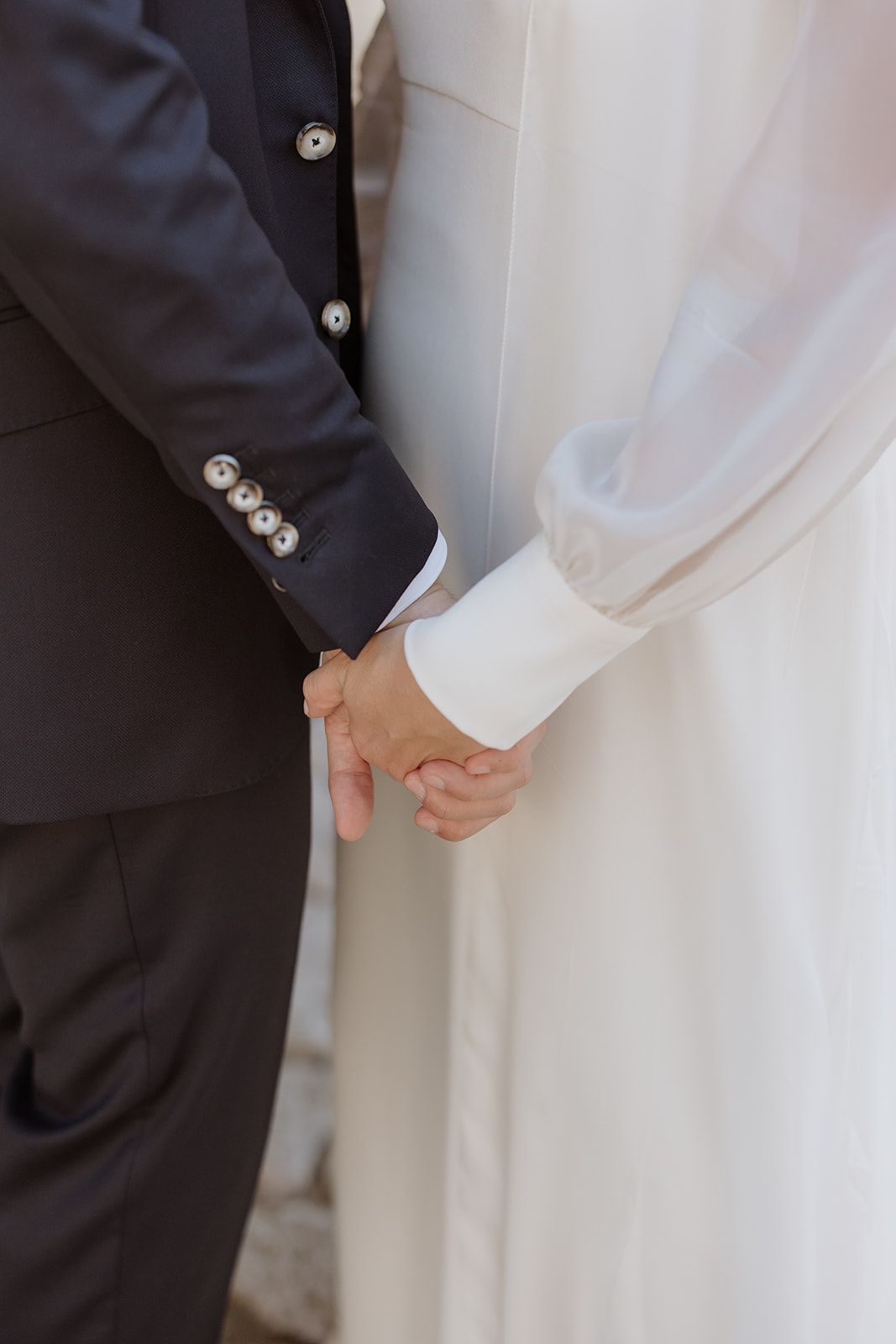 Close-up of joined hands between a groom in dark suit and bride in white dress.