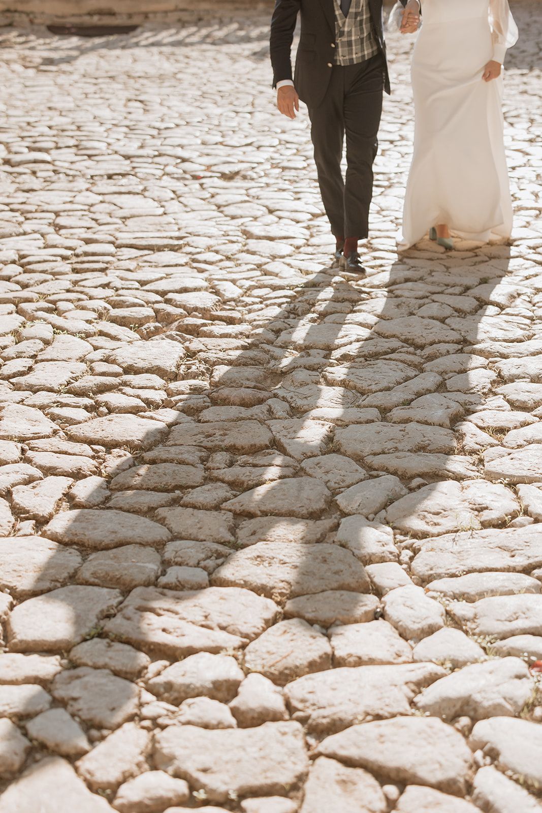 Wedding couple walking on cobblestone path casting long shadows.